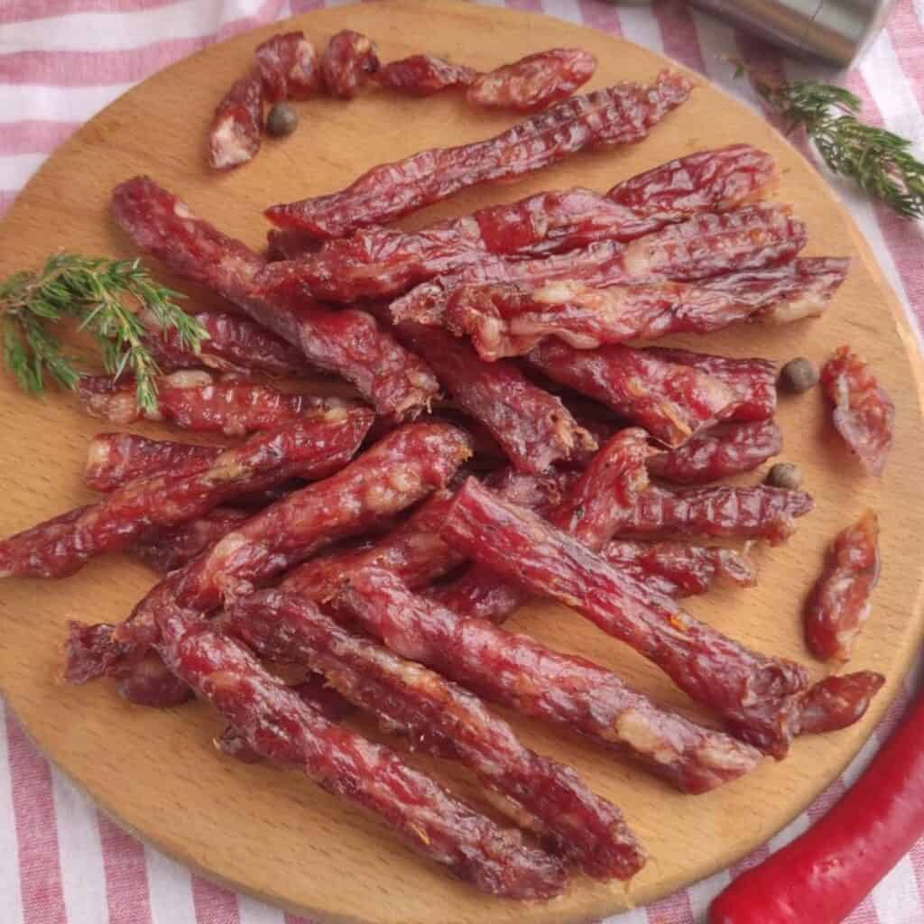 A round wooden board holds several pieces of thin, dried kabanos pork jerky arranged in a pile, garnished with a sprig of fresh herbs and a few whole peppercorns. The background features a red and white striped cloth.