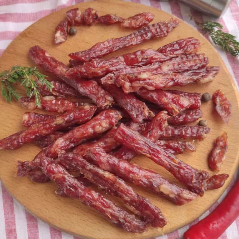 A round wooden board holds several pieces of thin, dried kabanos pork jerky arranged in a pile, garnished with a sprig of fresh herbs and a few whole peppercorns. The background features a red and white striped cloth.