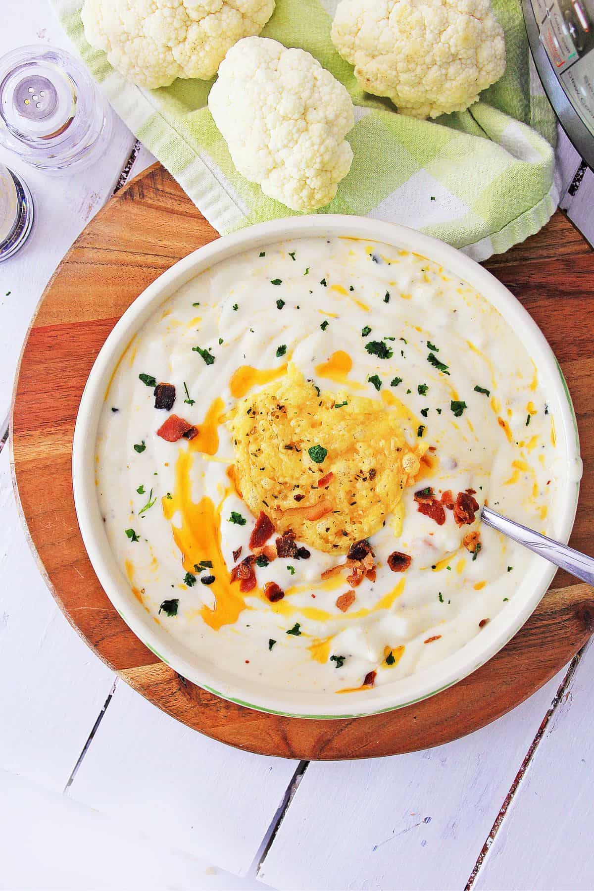 A bowl of creamy Cauliflower Parmesan Soup garnished with cheese, bacon pieces, and chopped herbs sits on a wooden board. Raw cauliflower and a green towel are in the background, while a spoon rests in the bowl.