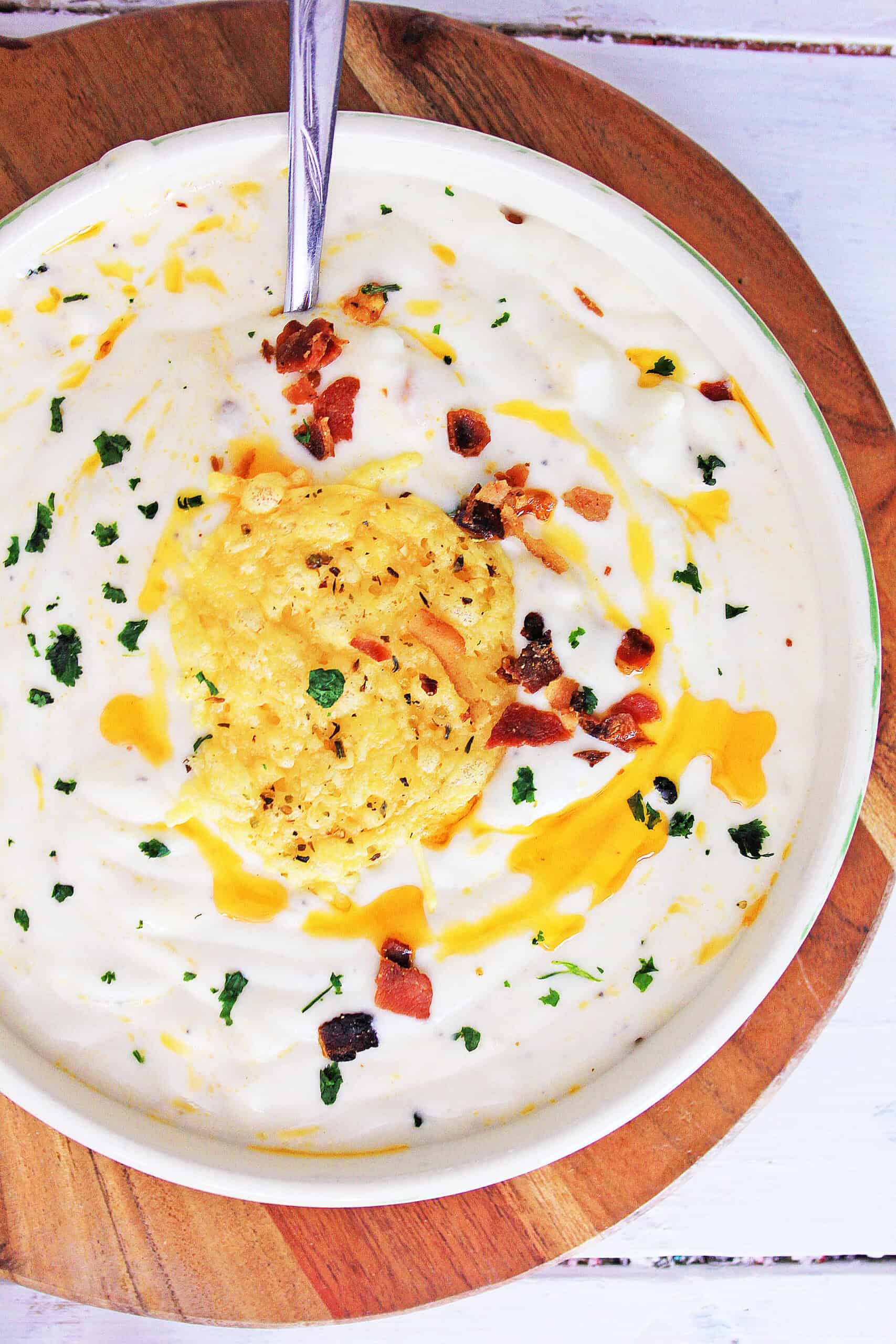 A bowl of creamy Cauliflower Parmesan Soup is topped with shredded cheese, crumbled bacon, chopped herbs, and a drizzle of yellow sauce, with a spoon resting inside. The bowl sits on a wooden board.