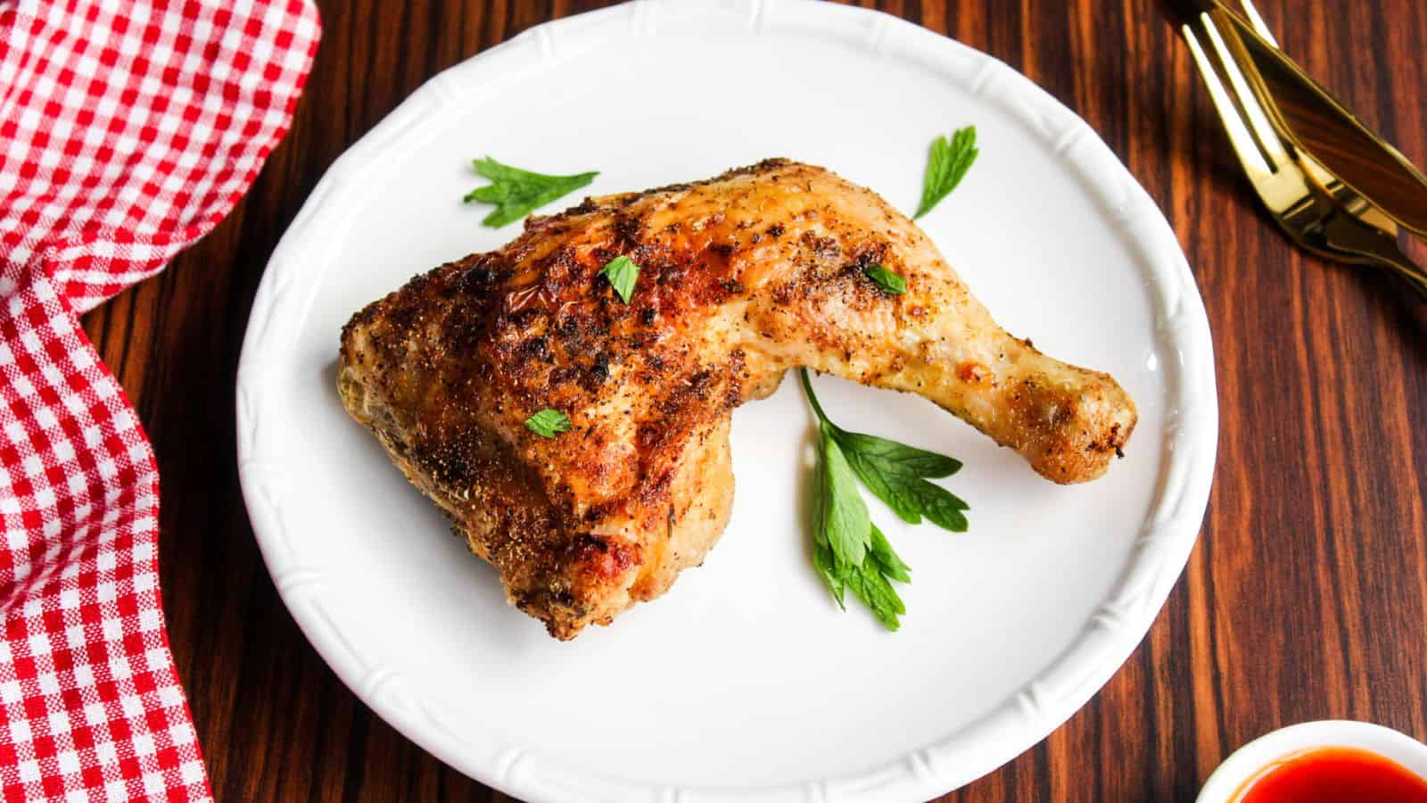 A cooked chicken leg quarter seasoned with herbs sits on a white plate, garnished with two sprigs of parsley. A red and white checkered napkin and gold utensils are on a wooden table nearby.