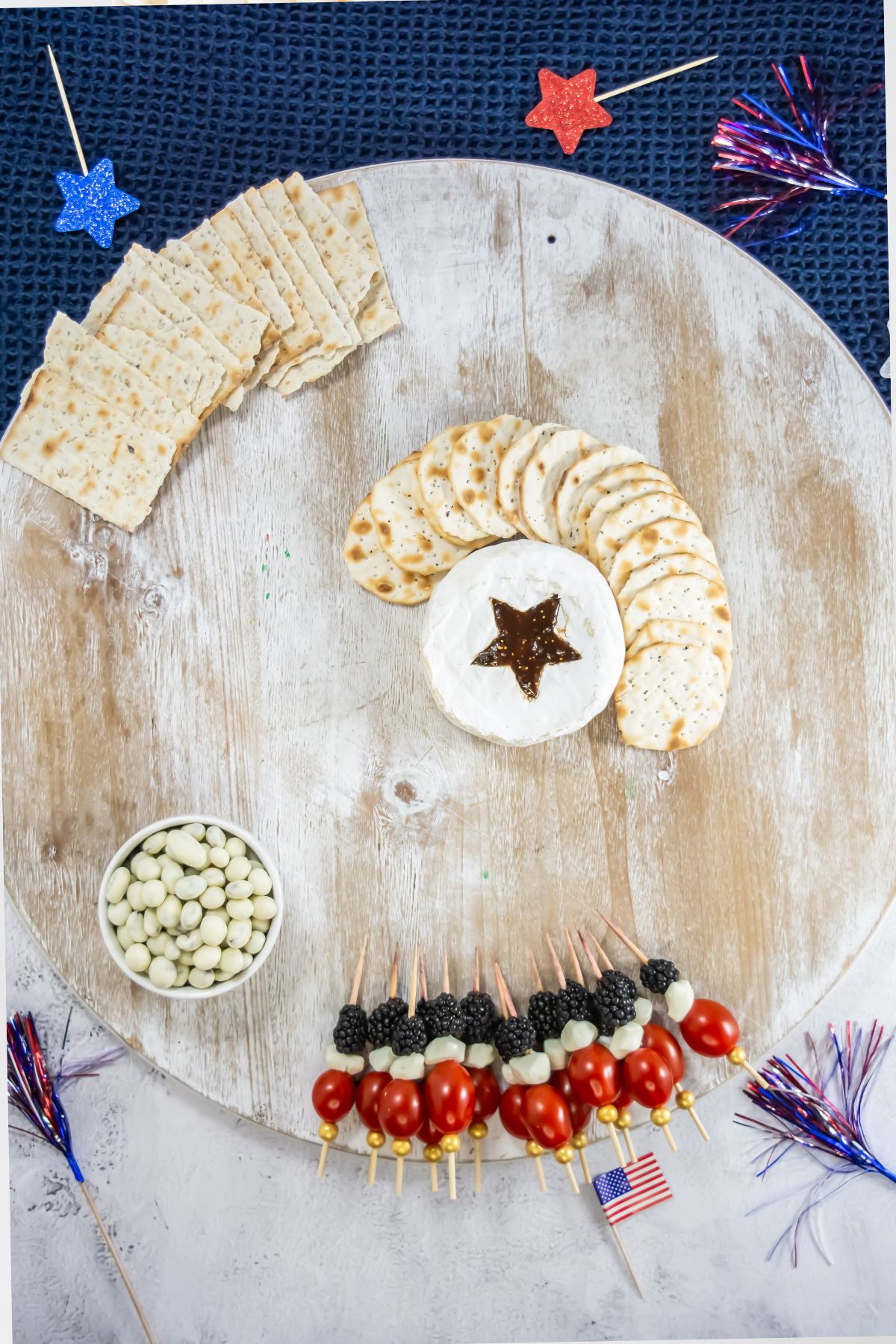 A round wooden board with rectangular and round crackers, a wheel of cheese with a star-shaped cutout in the center, a bowl of white snacks, and skewers of tomatoes, cheese, and berries. Red, white, and blue decorations are around the board.