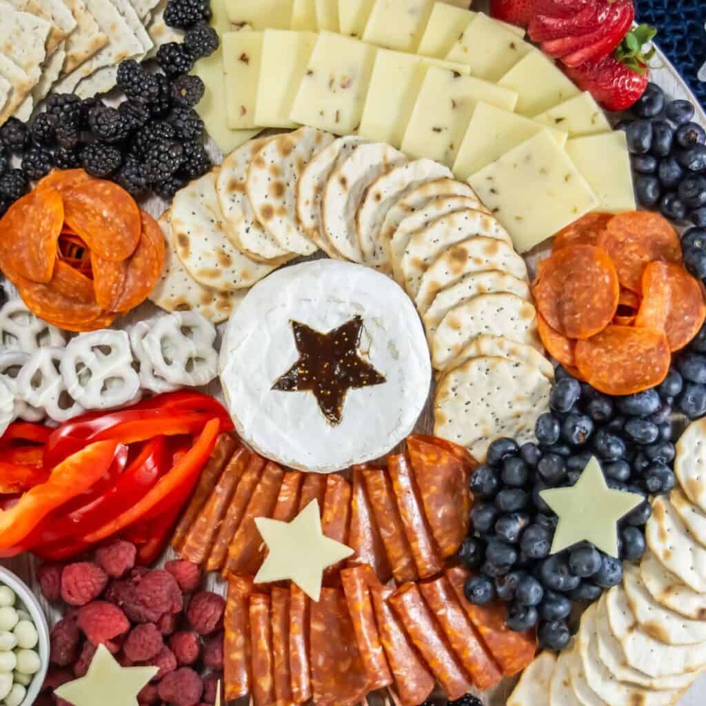 A charcuterie board with sliced cheese, crackers, pepperoni, salami, blackberries, blueberries, raspberries, strawberries, red bell pepper slices, white chocolate pretzels, and a round brie topped with a star-shaped jam center.