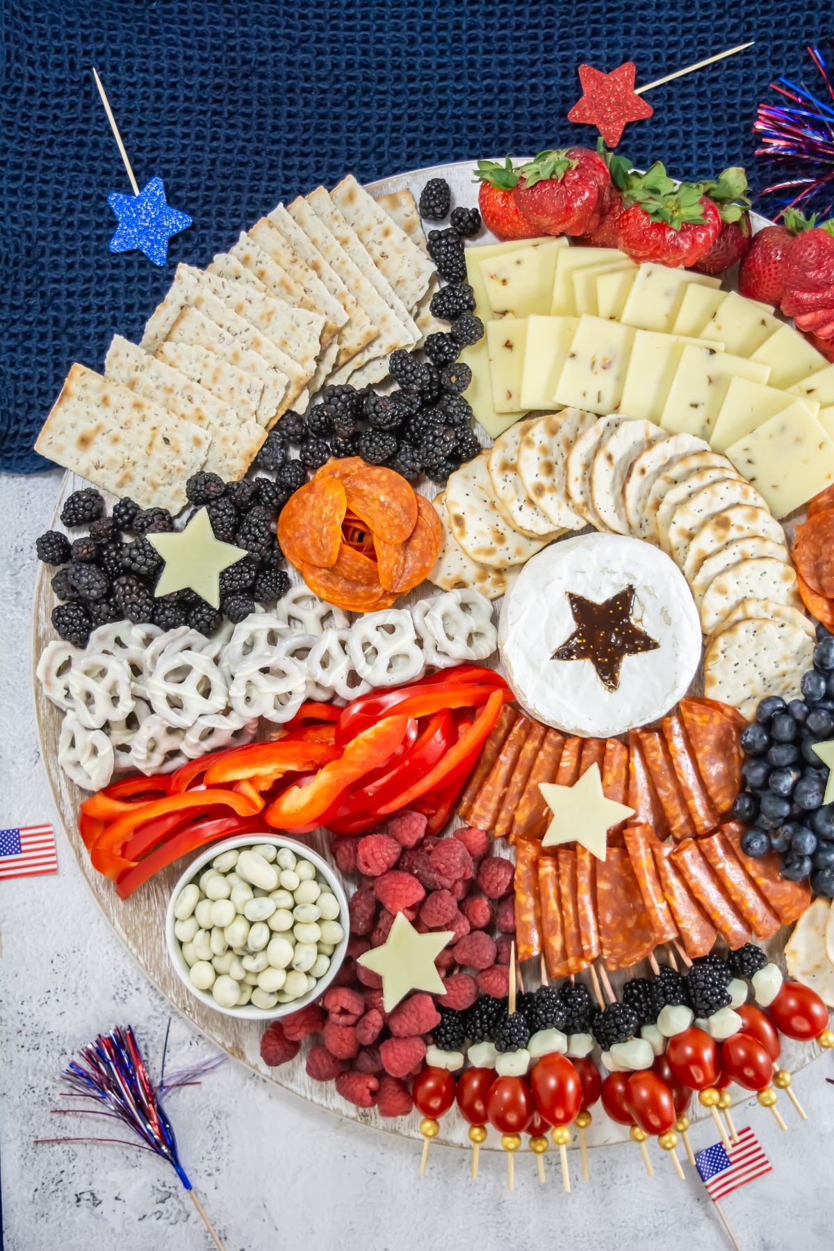 A round charcuterie board with assorted cheeses, crackers, sliced meats, berries, yogurt-covered pretzels, and star-shaped cheese pieces. Small American flags and star decorations are placed around the board.