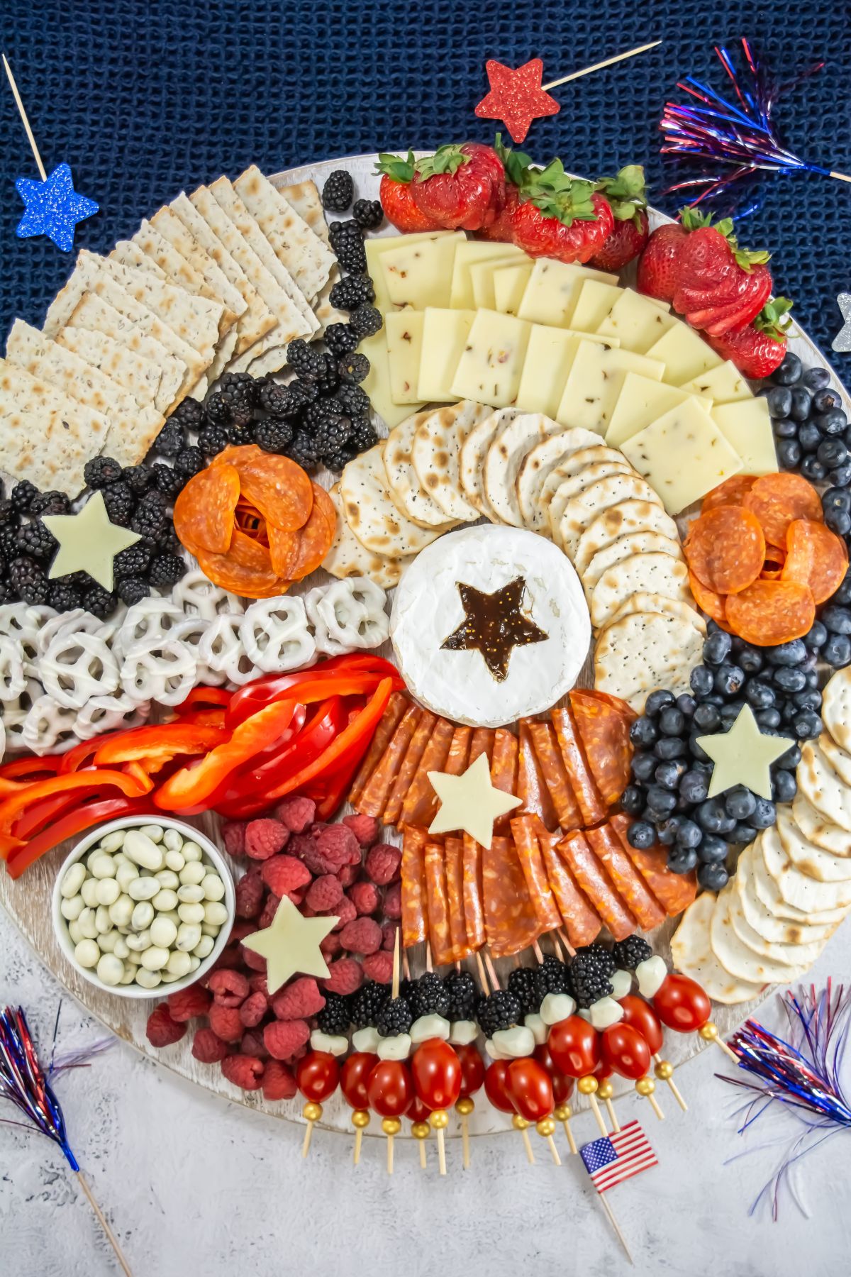 A round charcuterie board with cheese, crackers, salami, pepperoni, berries, pretzels, and vegetables, arranged in a red, white, and blue patriotic pattern with star-shaped cheese and American flag decorations.