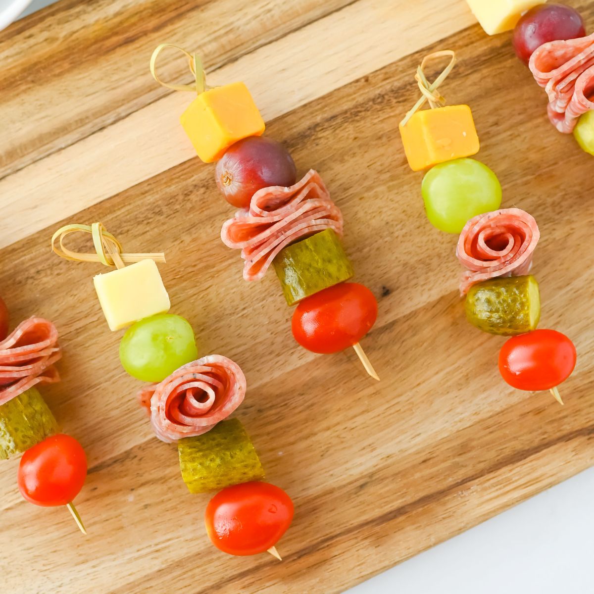 Skewers on a wooden board, each with a cherry tomato, pickle slice, folded salami, green grape, cheese cube, and red grape neatly arranged in a row.