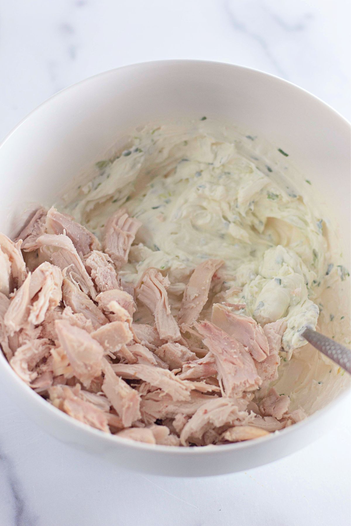 A white mixing bowl containing shredded cooked chicken and a creamy mixture with herbs, partially stirred together, with a metal spoon in the bowl. The bowl is on a white countertop.