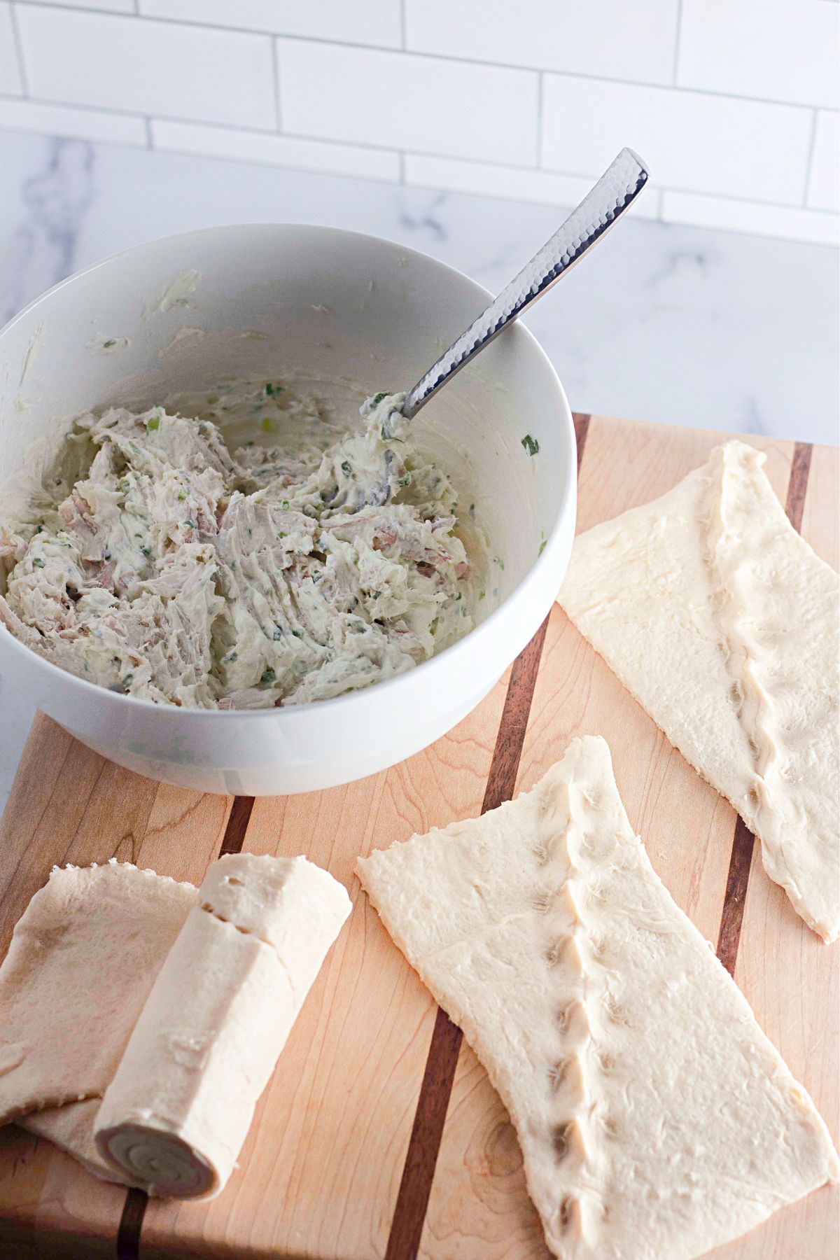 A white bowl with a creamy mixture and a spoon inside sits on a wooden cutting board next to unrolled crescent dough triangles and a partially unrolled dough canister. White subway tile background.