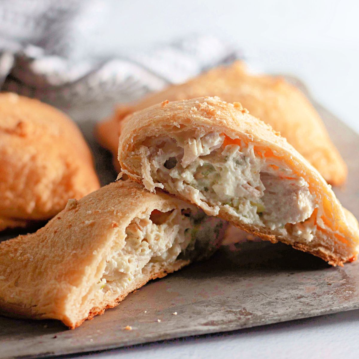 A golden-brown baked pastry is cut in half, revealing a creamy filling with visible bits of what appear to be chicken and vegetables. Other whole pastries are in the background on a baking sheet.