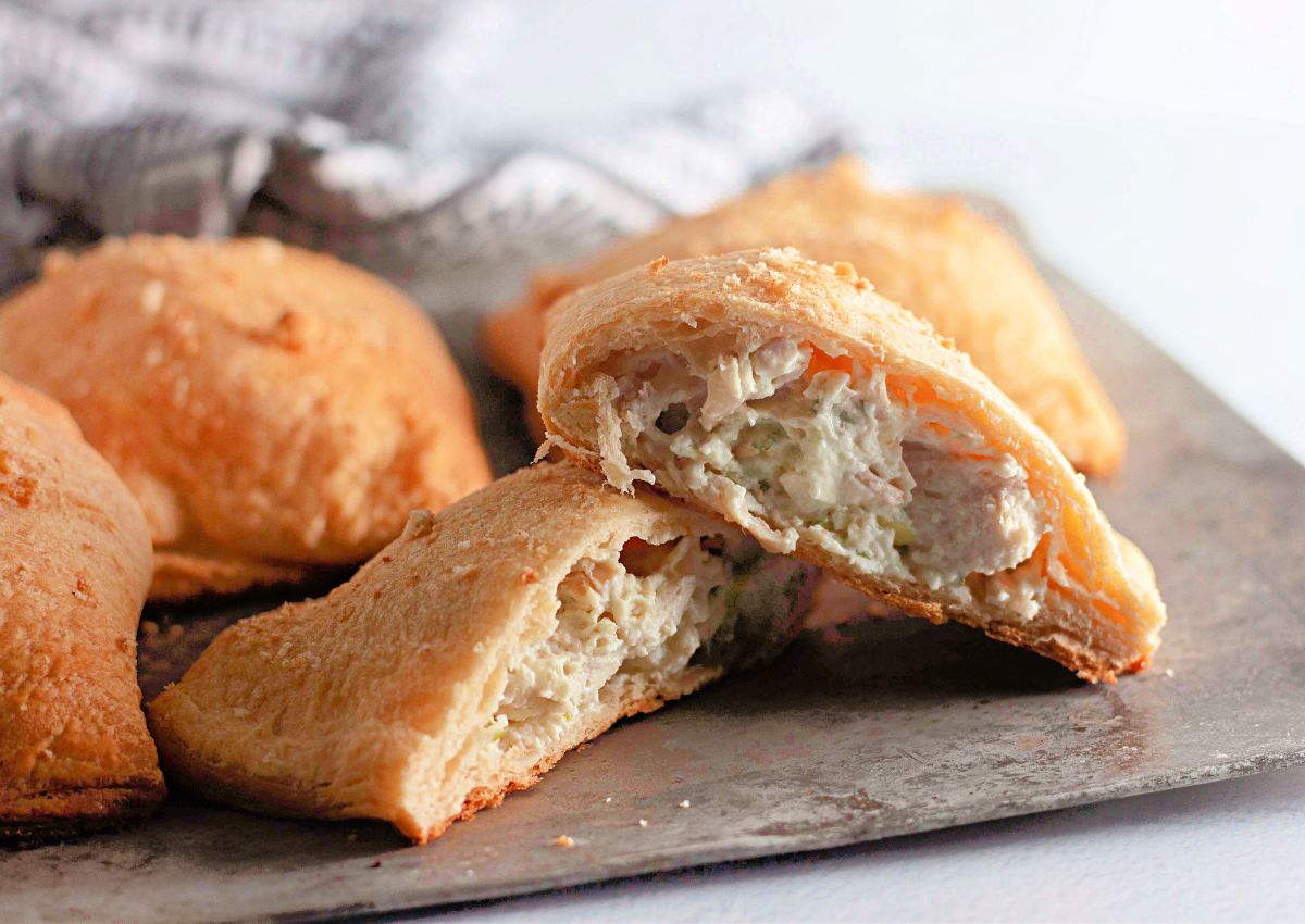A close-up of a baked pastry cut in half to show a creamy, chicken and vegetable filling. Other whole pastries are visible in the background on a metal baking sheet.
