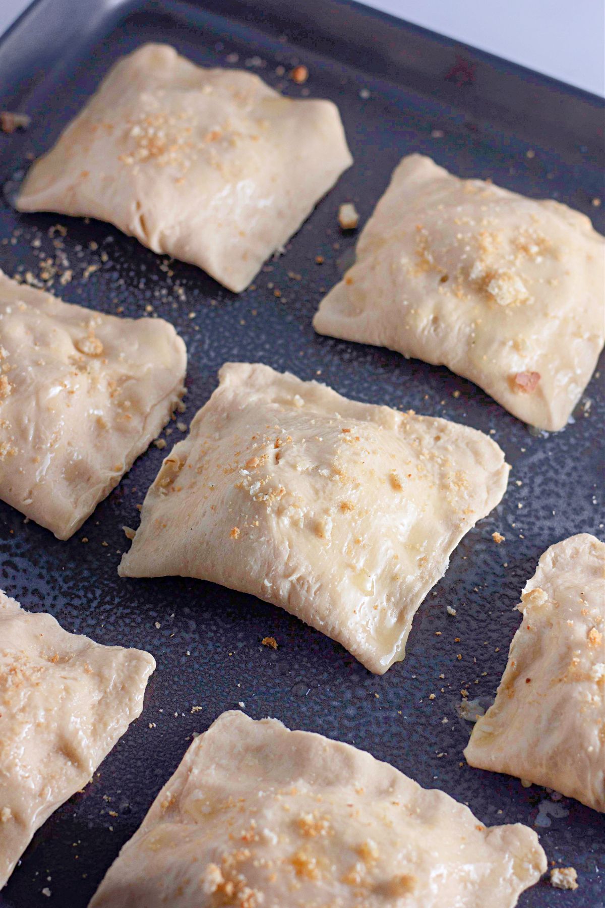 Unbaked, square ravioli pieces are arranged on a dark baking sheet. The dough appears soft and lightly dusted with flour or crumbs, ready to be cooked.