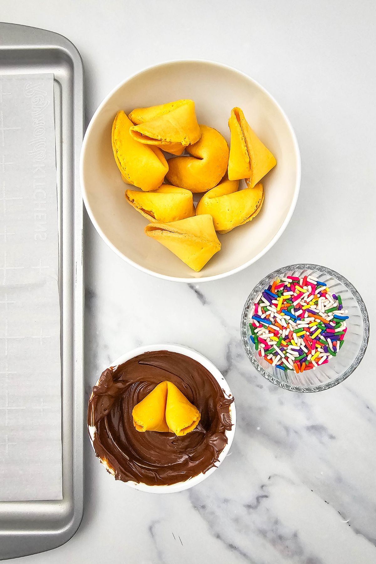 A bowl of fortune cookies, a small glass bowl of rainbow sprinkles, and a bowl with melted chocolate holding a dipped fortune cookie sit on a white marble surface next to a baking sheet lined with parchment paper.