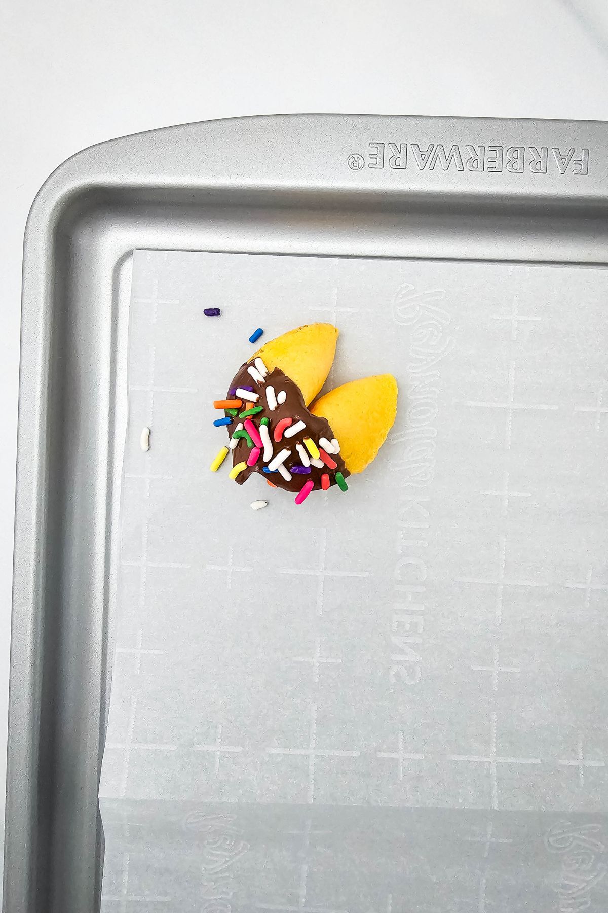 A fortune cookie dipped in chocolate and covered with colorful sprinkles sits on the corner of a parchment-lined baking sheet.