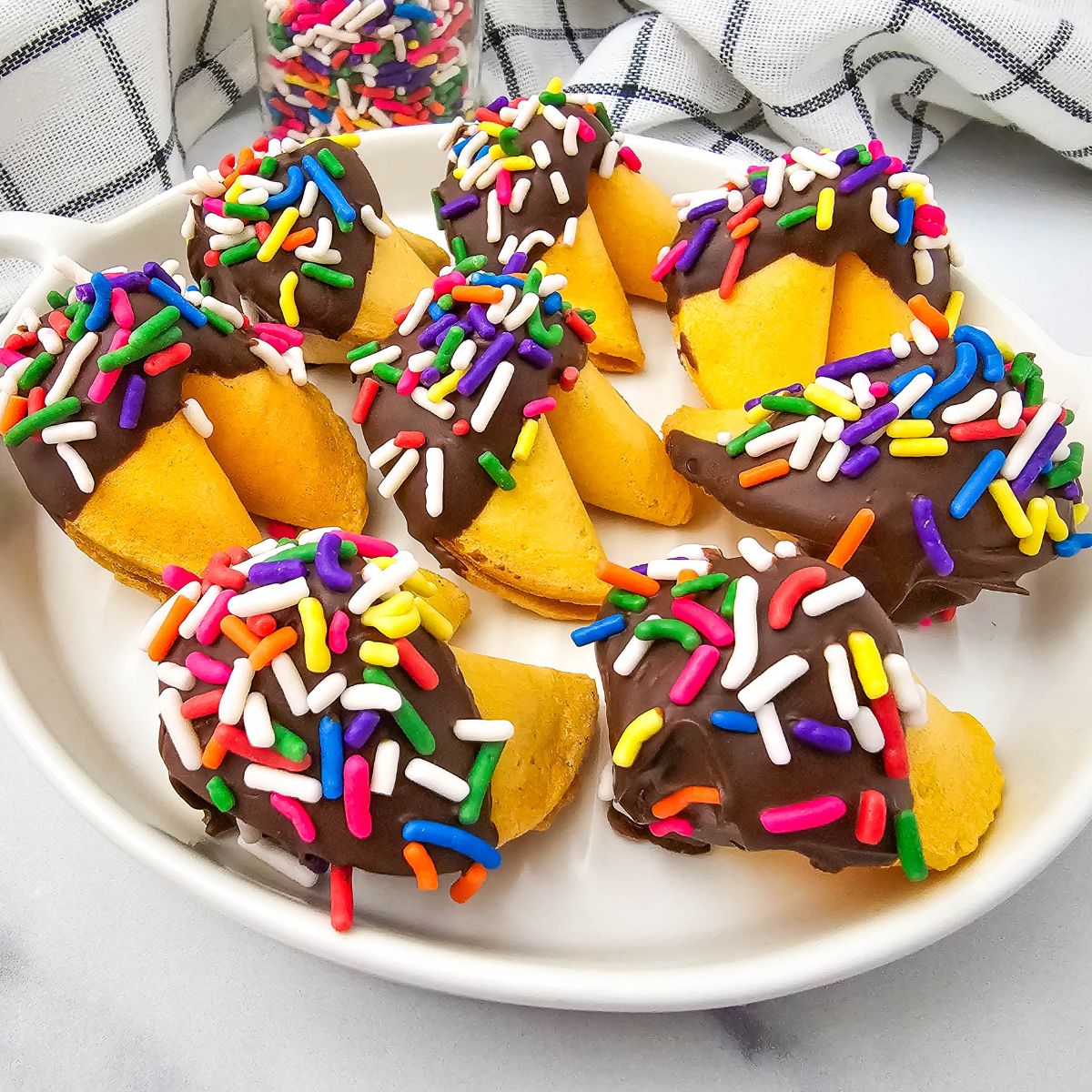 A white plate holds fortune cookies dipped in chocolate and topped with colorful sprinkles. A jar of sprinkles and a checkered cloth are in the background.