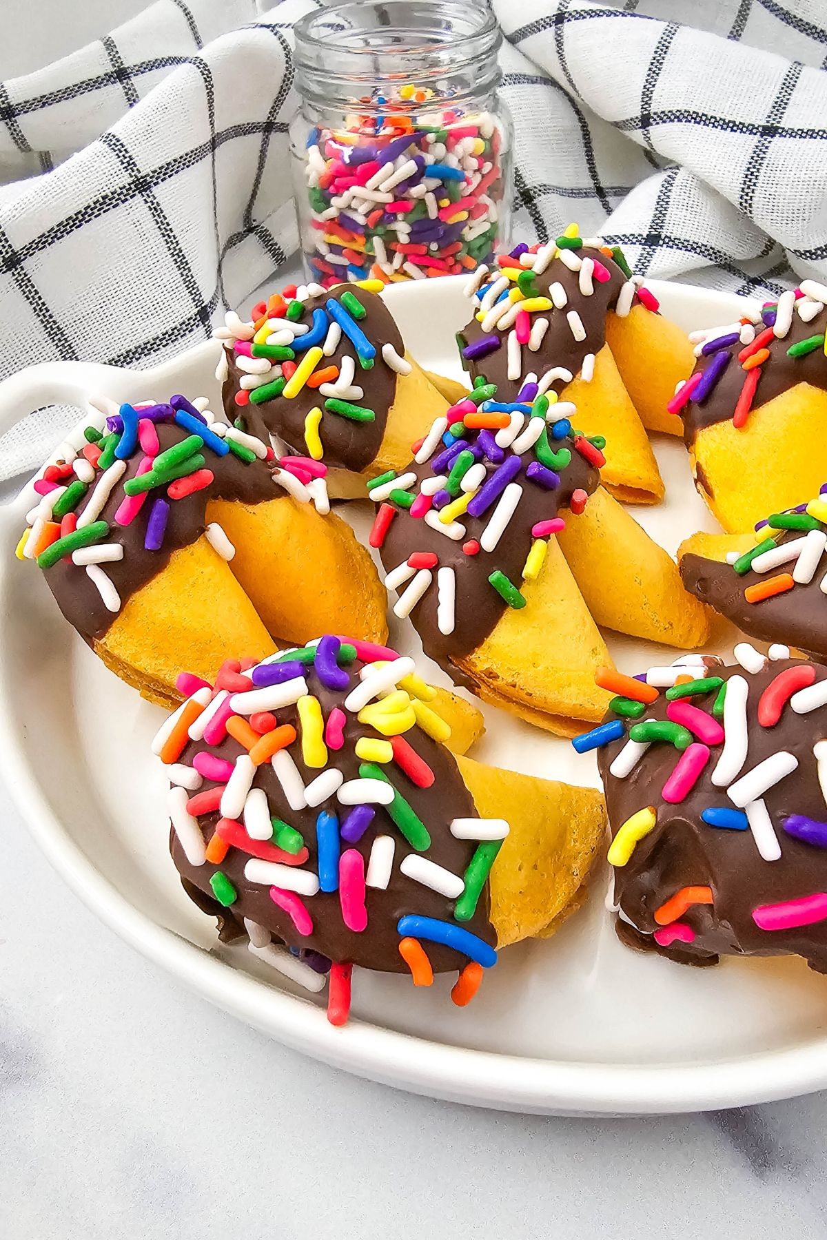 A plate of fortune cookies partially dipped in chocolate and covered with colorful sprinkles is displayed in the foreground, with a jar of extra sprinkles and a white checkered towel in the background.