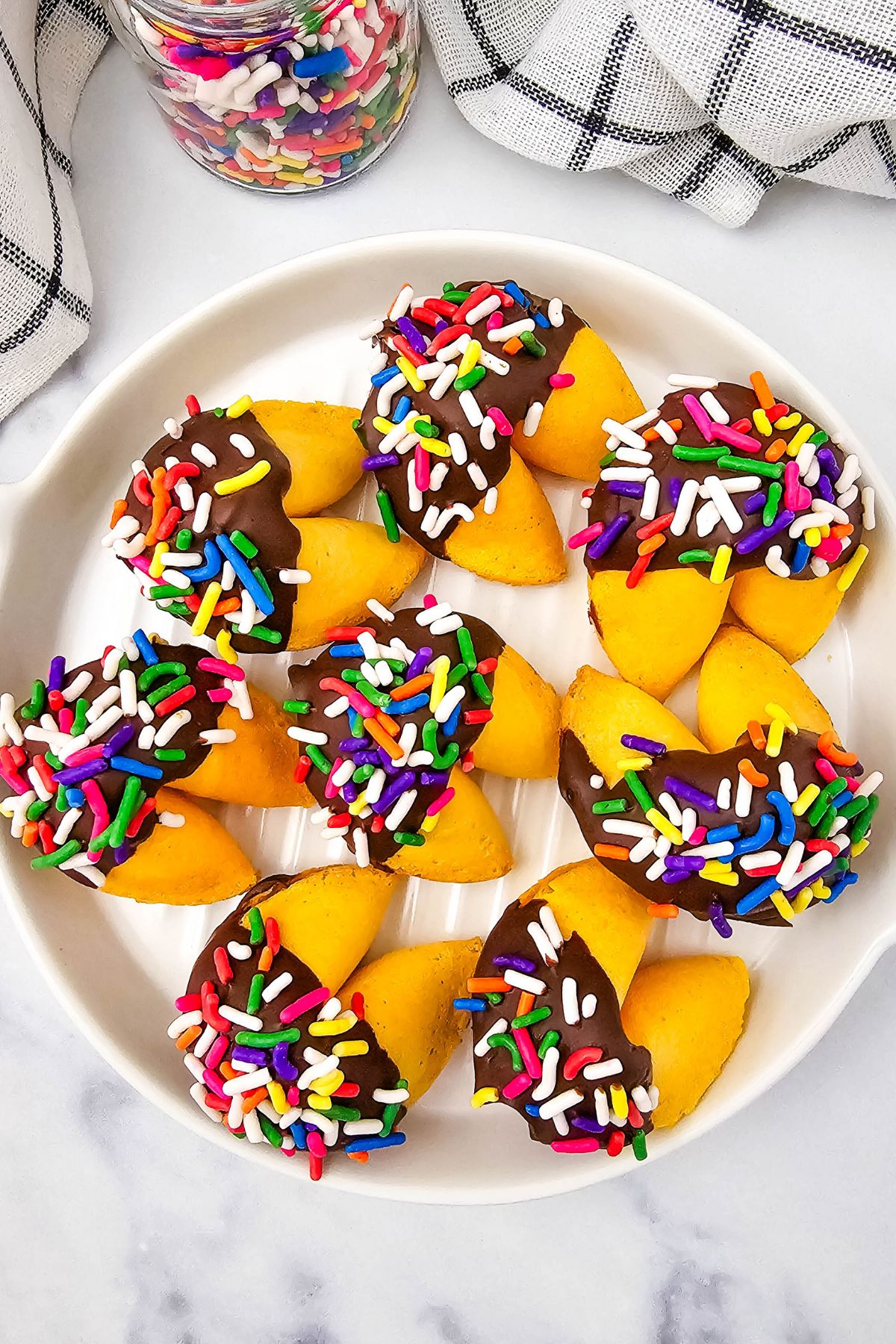 A white plate holds yellow fortune cookies dipped in chocolate and covered with colorful sprinkles. A jar of sprinkles and a black-and-white checkered cloth are visible in the background.