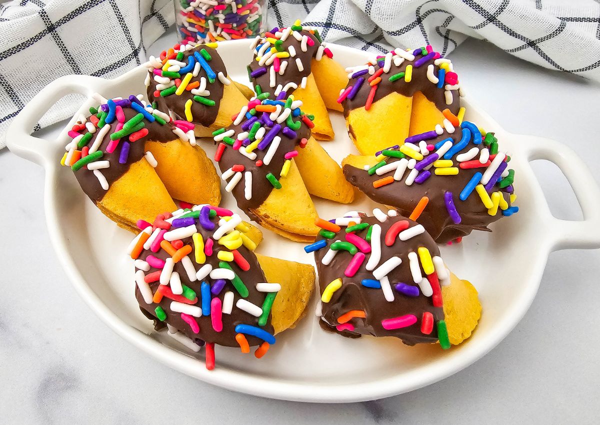 A white dish holds fortune cookies dipped in chocolate and topped with colorful rainbow sprinkles. A black and white checkered towel is in the background.