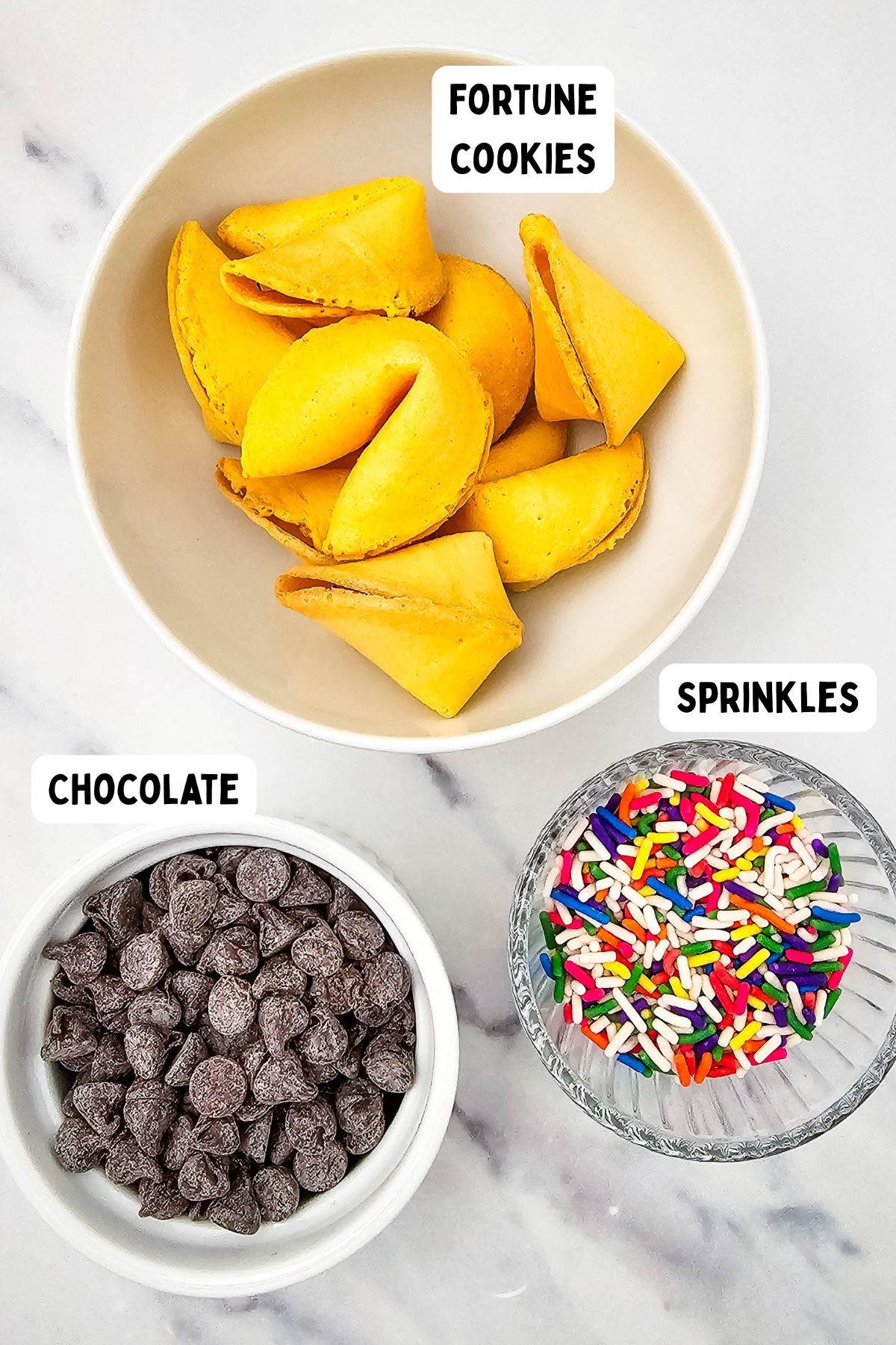 A bowl of fortune cookies, a bowl of chocolate chips, and a bowl of rainbow sprinkles are arranged on a white marble surface. Each bowl is labeled: fortune cookies, chocolate, and sprinkles.