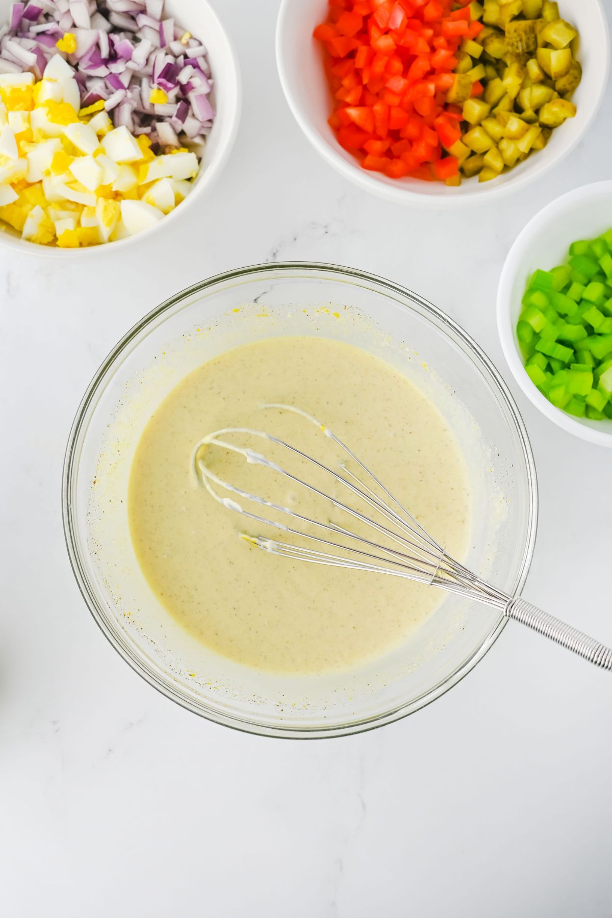 A glass bowl with a whisk inside containing a creamy dressing sits on a white surface. Surrounding it are bowls filled with chopped red bell pepper, celery, pickles, eggs, and red onion.