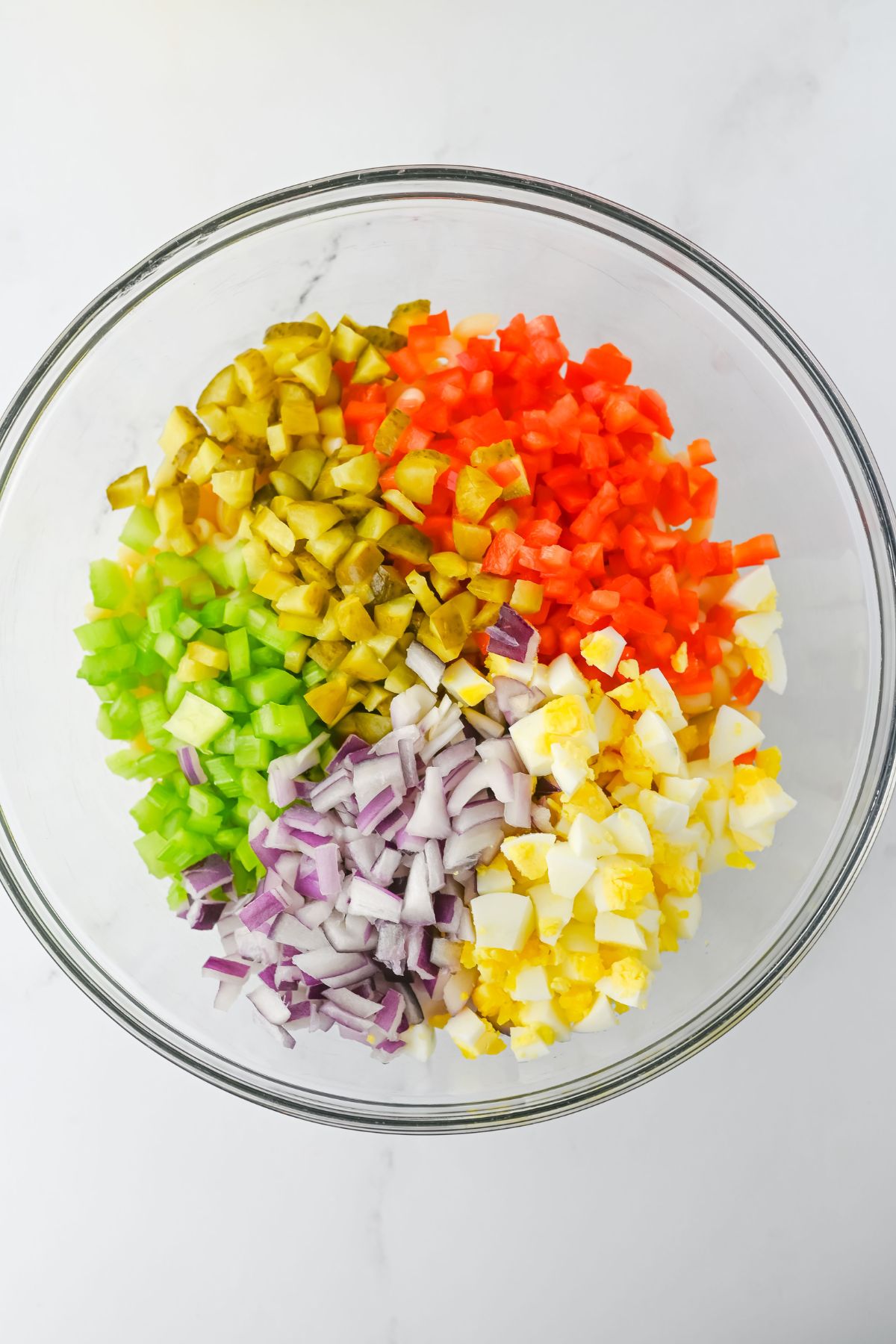 A clear glass bowl containing separate piles of chopped green bell pepper, pickles, red bell pepper, white onion, red onion, and hard-boiled egg on a white surface.