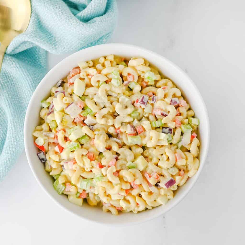A white bowl filled with macaroni salad containing elbow pasta, chopped celery, red onion, and red bell pepper on a white surface beside a light blue napkin.