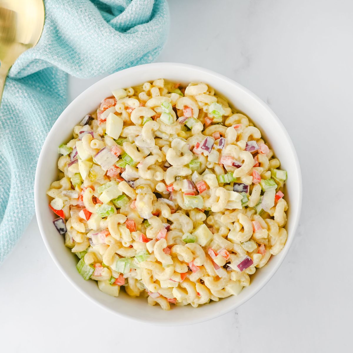 A white bowl filled with macaroni salad containing elbow pasta, chopped celery, red onion, and red bell pepper on a white surface beside a light blue napkin.