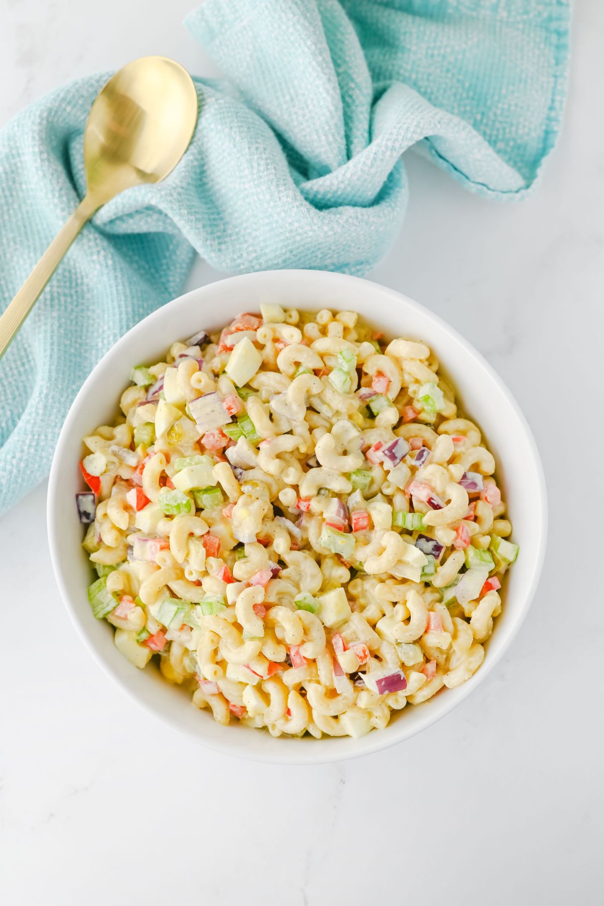 A white bowl filled with macaroni salad containing diced vegetables sits on a white surface next to a light blue textured cloth and a gold-colored spoon.