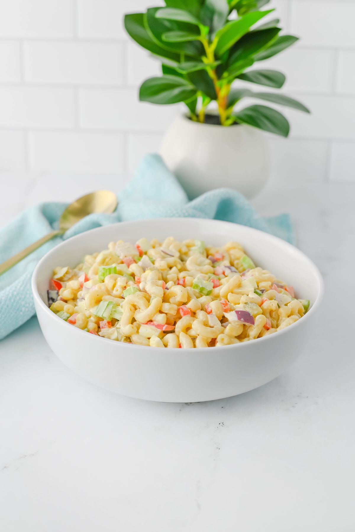A white bowl filled with macaroni salad containing diced vegetables sits on a white surface. A gold spoon, a light blue cloth, and a small potted plant are in the background.
