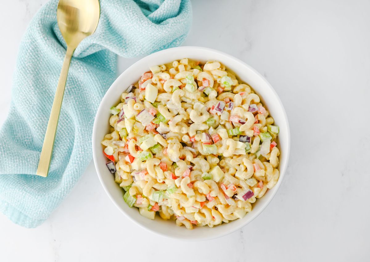A white bowl filled with macaroni salad containing elbow pasta, diced red onion, celery, and red bell pepper. A gold spoon and a light blue cloth are placed beside the bowl on a white surface.