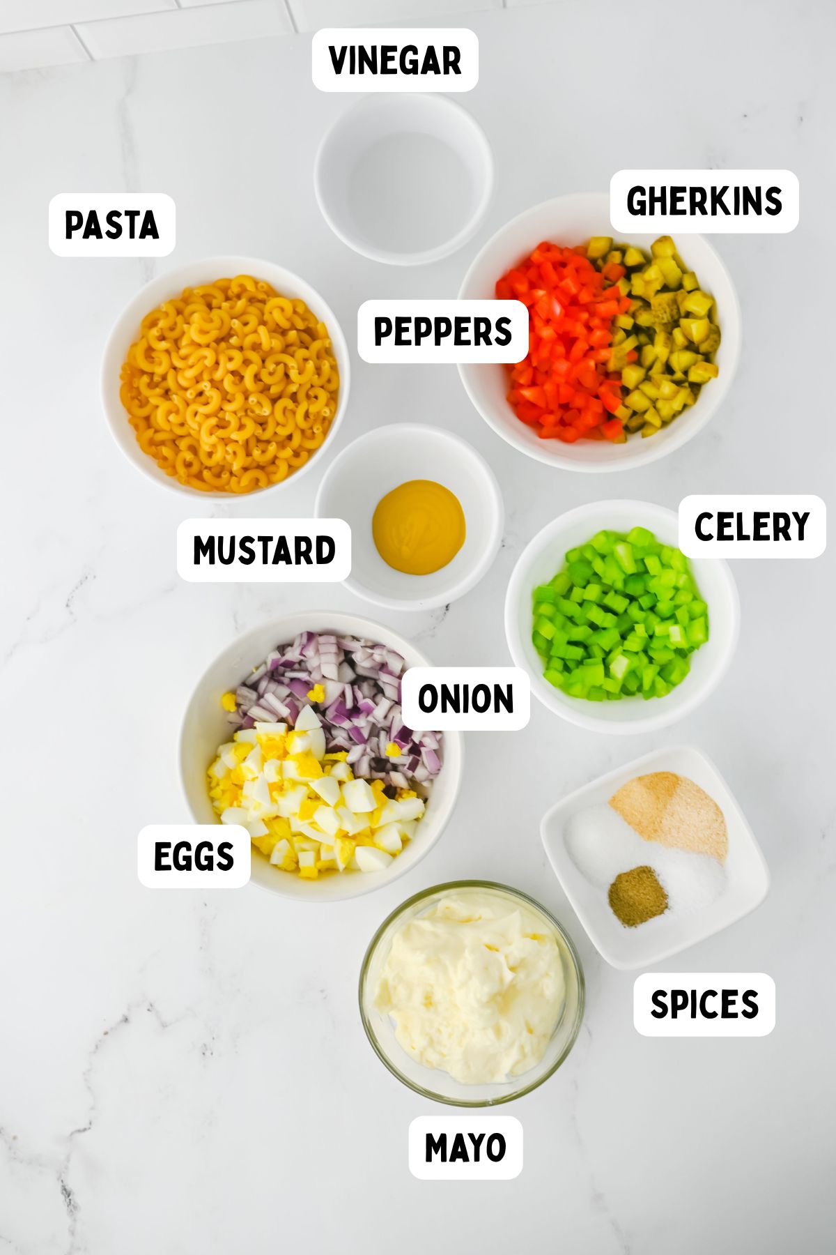 Nine bowls on a white surface contain labeled ingredients for pasta salad: pasta, vinegar, gherkins, peppers, mustard, celery, onion, eggs, mayo, and a bowl of spices.