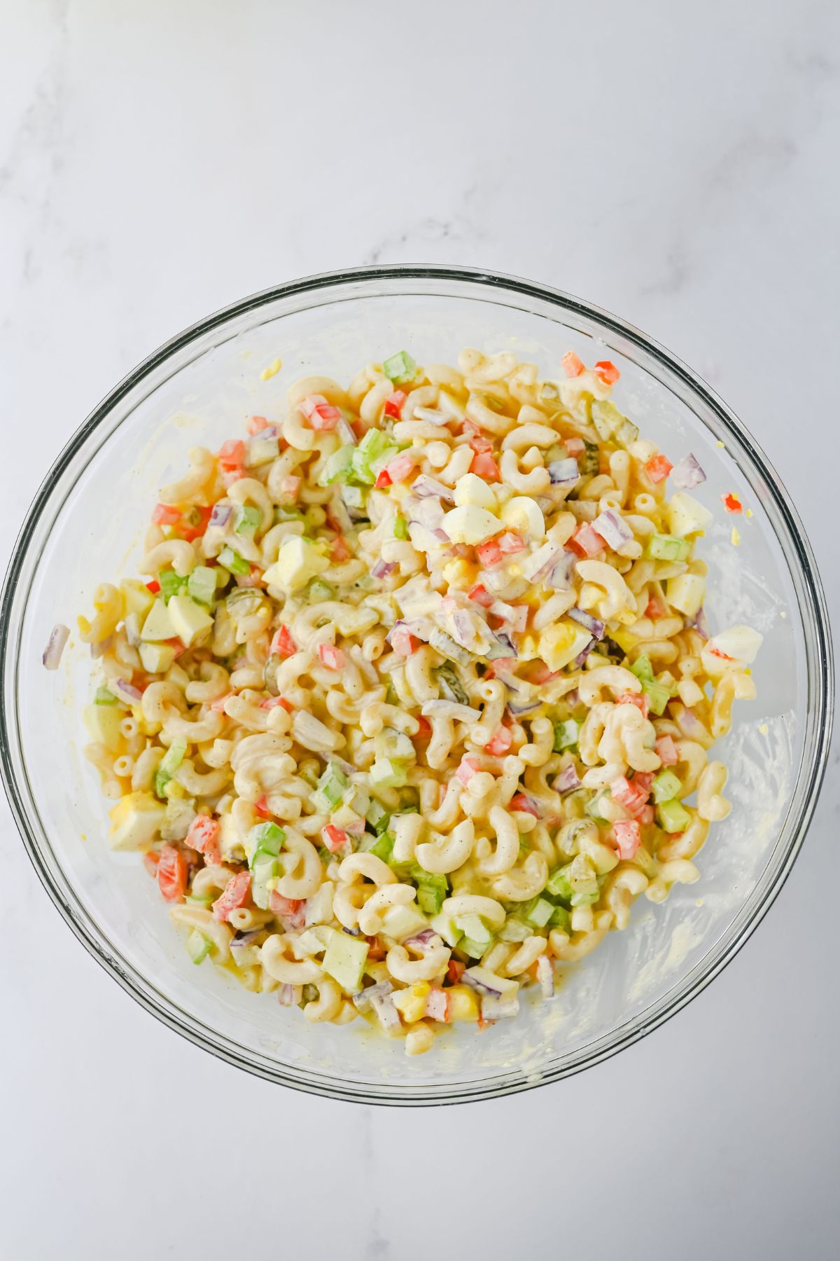 A clear glass bowl filled with macaroni salad containing elbow pasta, diced red onion, celery, pickles, and red bell pepper, mixed with a creamy dressing, on a white marble surface.