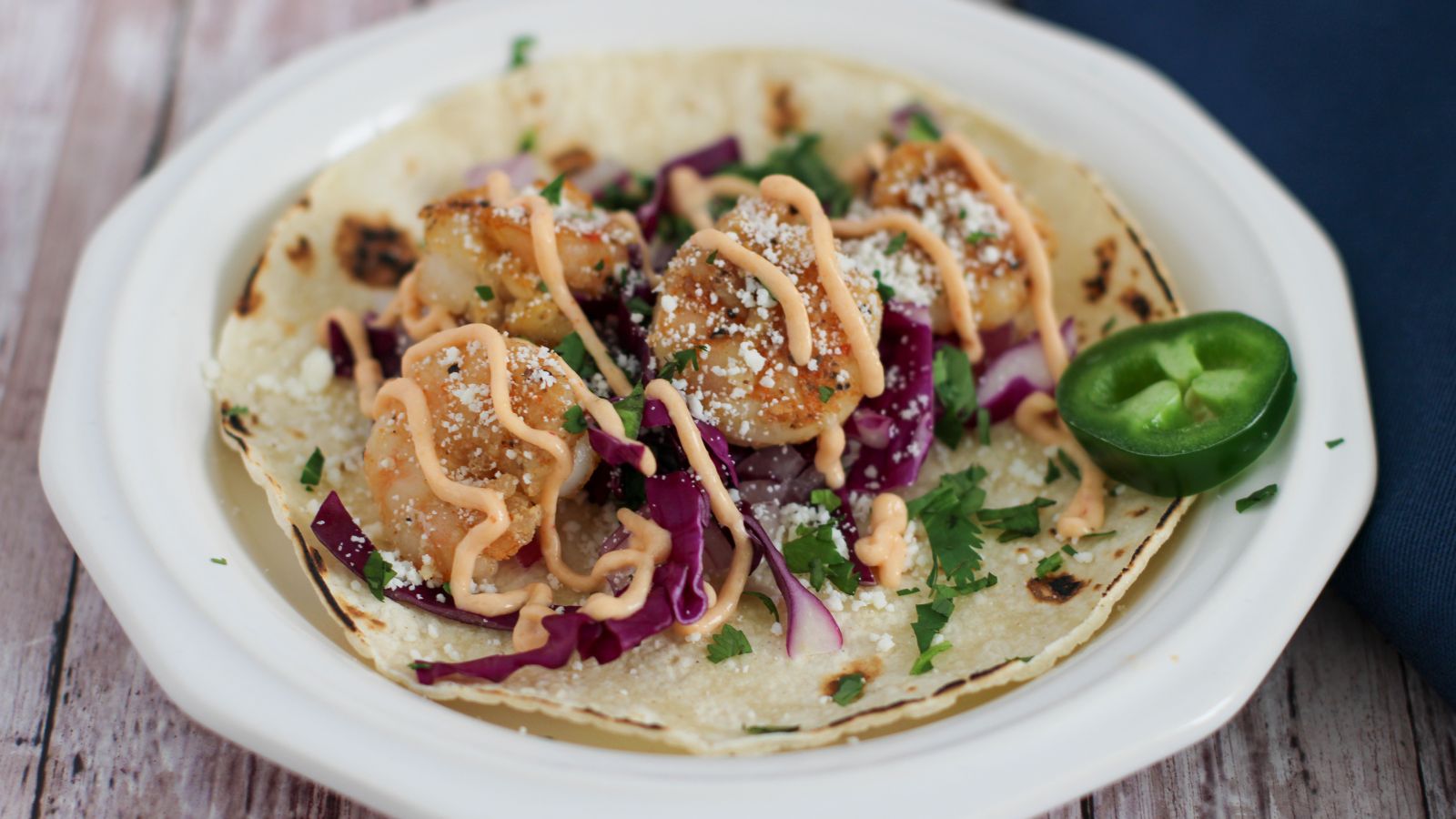 Shrimp taco with cabbage, onion, cilantro, sauce, cheese, and a lime wedge on a corn tortilla.