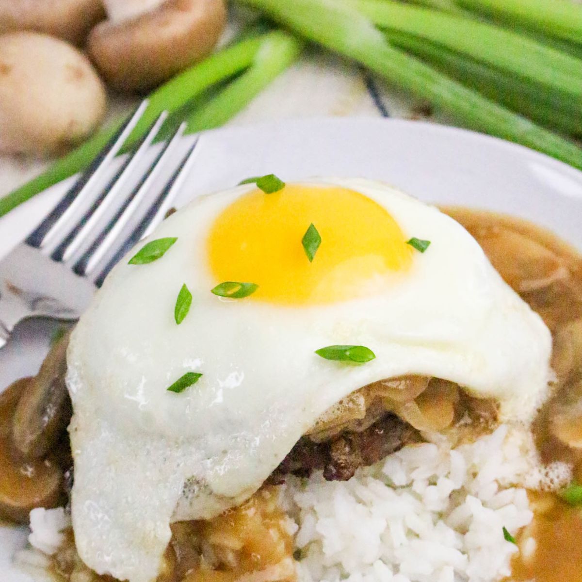 A plate of white rice topped with a hamburger patty, brown gravy with mushrooms, and a sunny-side-up egg, garnished with chopped green onions. A fork and green onions are in the background.