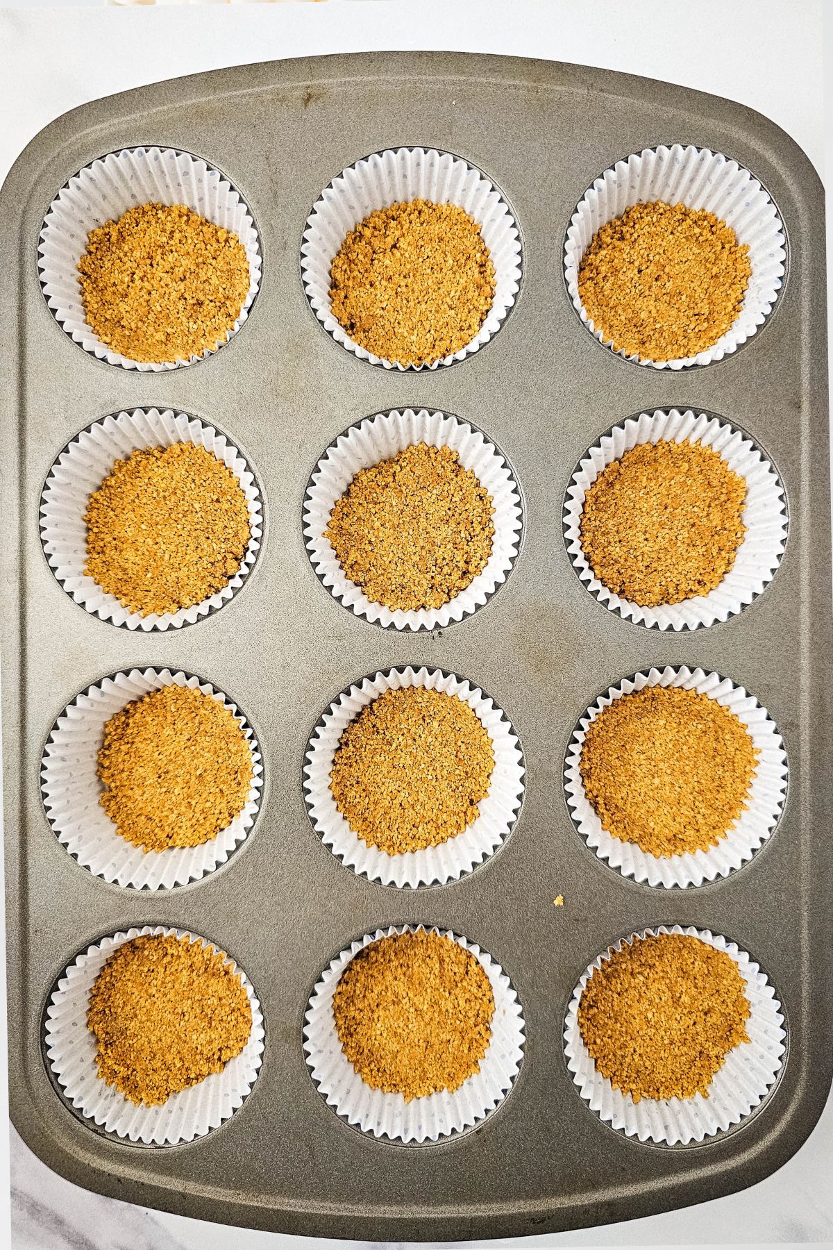A muffin tin holds twelve paper liners, each filled with a layer of crushed graham cracker crumbs, evenly distributed to form the base for individual desserts.