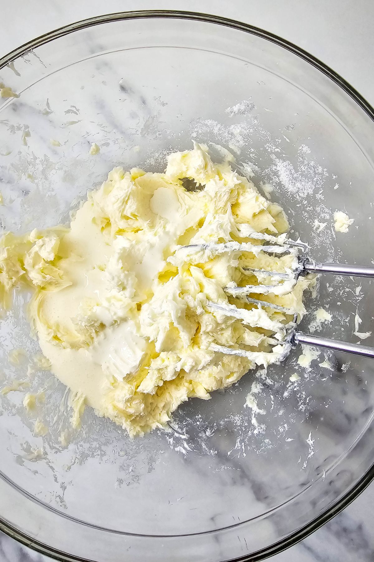 A clear glass bowl containing creamed butter and sugar being mixed with metal beaters on a marble surface.