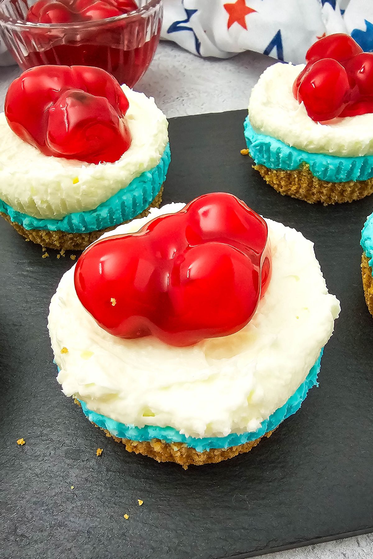 Mini cheesecakes with a crumb crust and white filling are topped with glossy red cherries. They are wrapped in blue cupcake liners and displayed on a dark slate surface. A white napkin with red and blue stars is in the background.