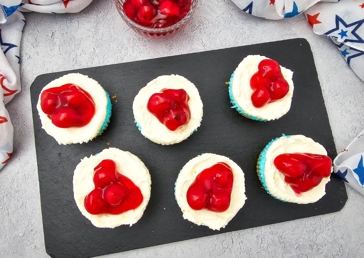 Six cupcakes with white frosting and cherry topping are arranged on a black rectangular slate. A bowl of extra cherry topping and a white star-patterned cloth are visible in the background.