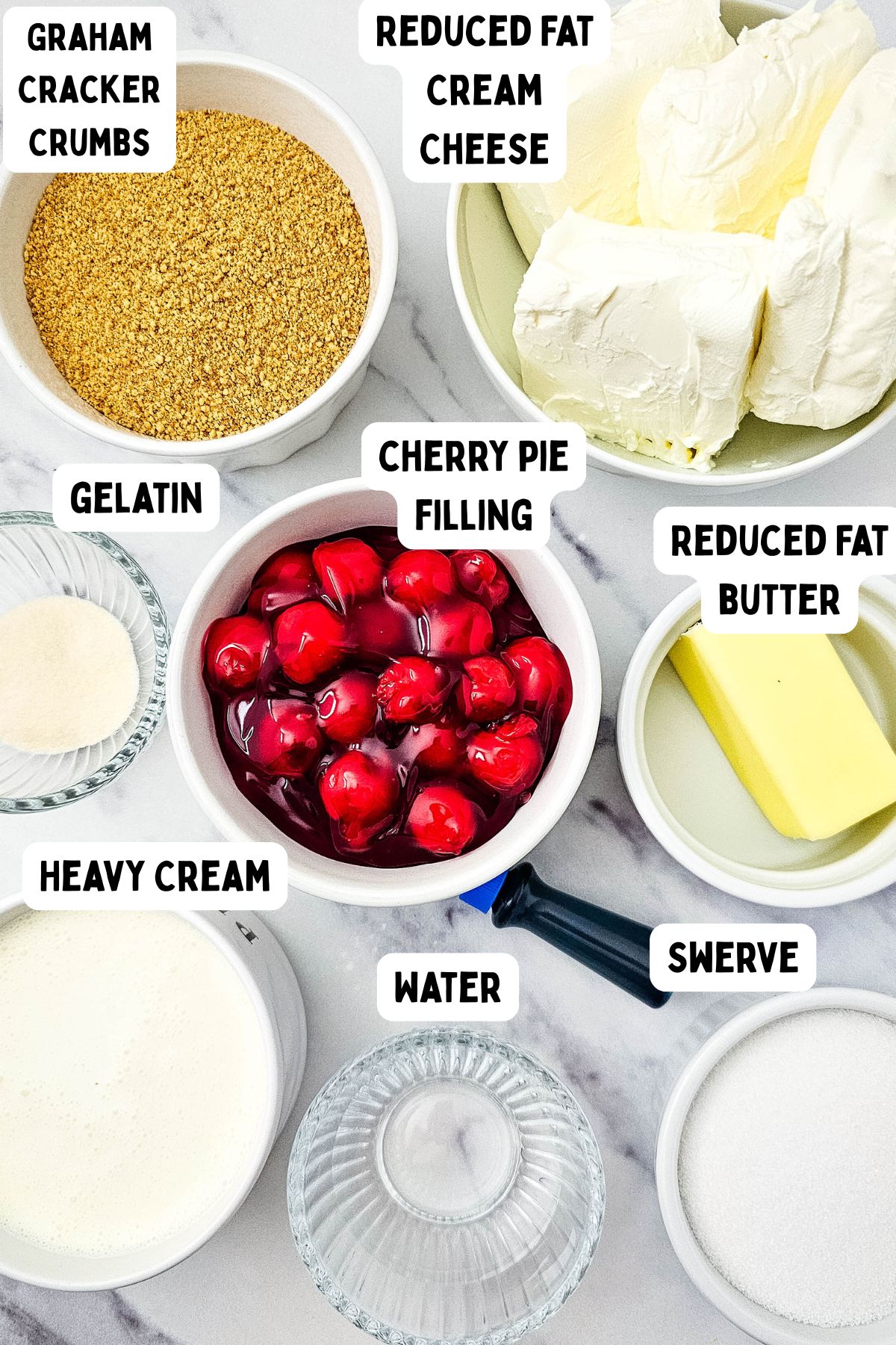 Top-down view of bowls and plates containing labeled ingredients: graham cracker crumbs, reduced fat cream cheese, gelatin, cherry pie filling, heavy cream, water, reduced fat butter, and Swerve, arranged on a white surface.
