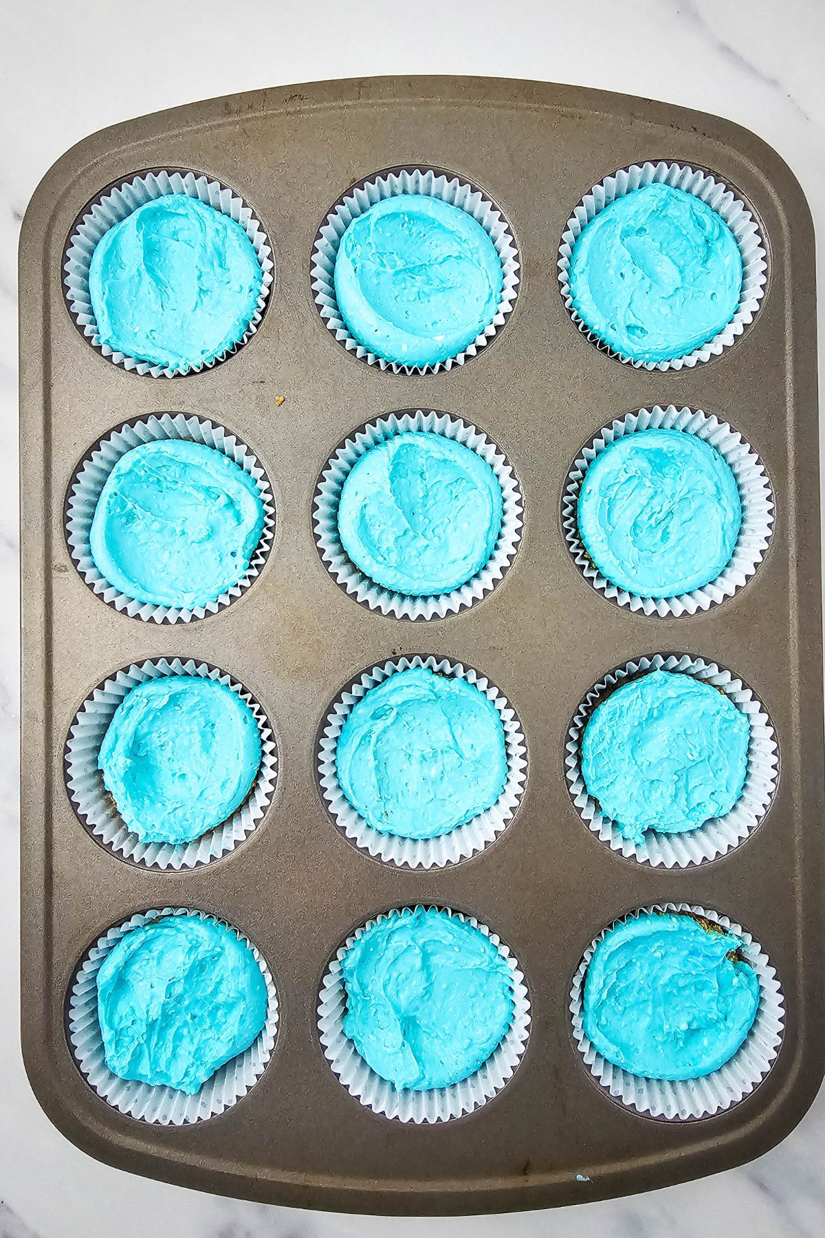 A muffin tin containing twelve cupcakes with bright blue frosting, each in a white paper liner, sits on a white countertop.