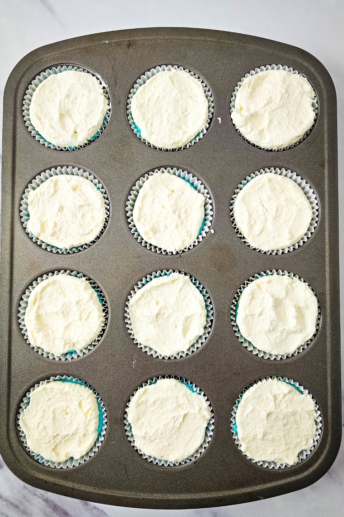 A metal muffin tin holds twelve paper cupcake liners, each filled with an even amount of unbaked batter. The tray is placed on a light-colored surface.