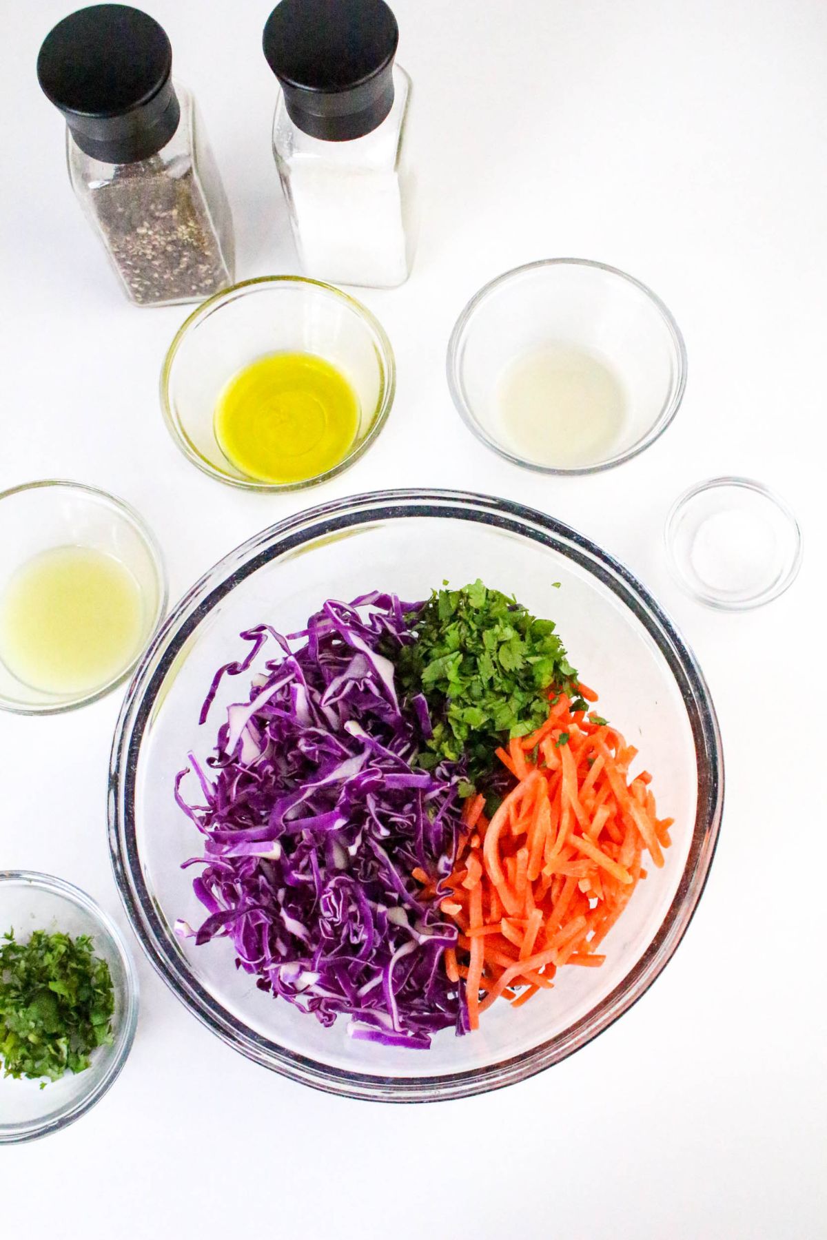 A glass bowl with shredded purple cabbage, chopped cilantro, and julienned carrots. Surrounding it are small bowls of olive oil, lemon juice, and white vinegar, plus salt and pepper shakers on a white surface.