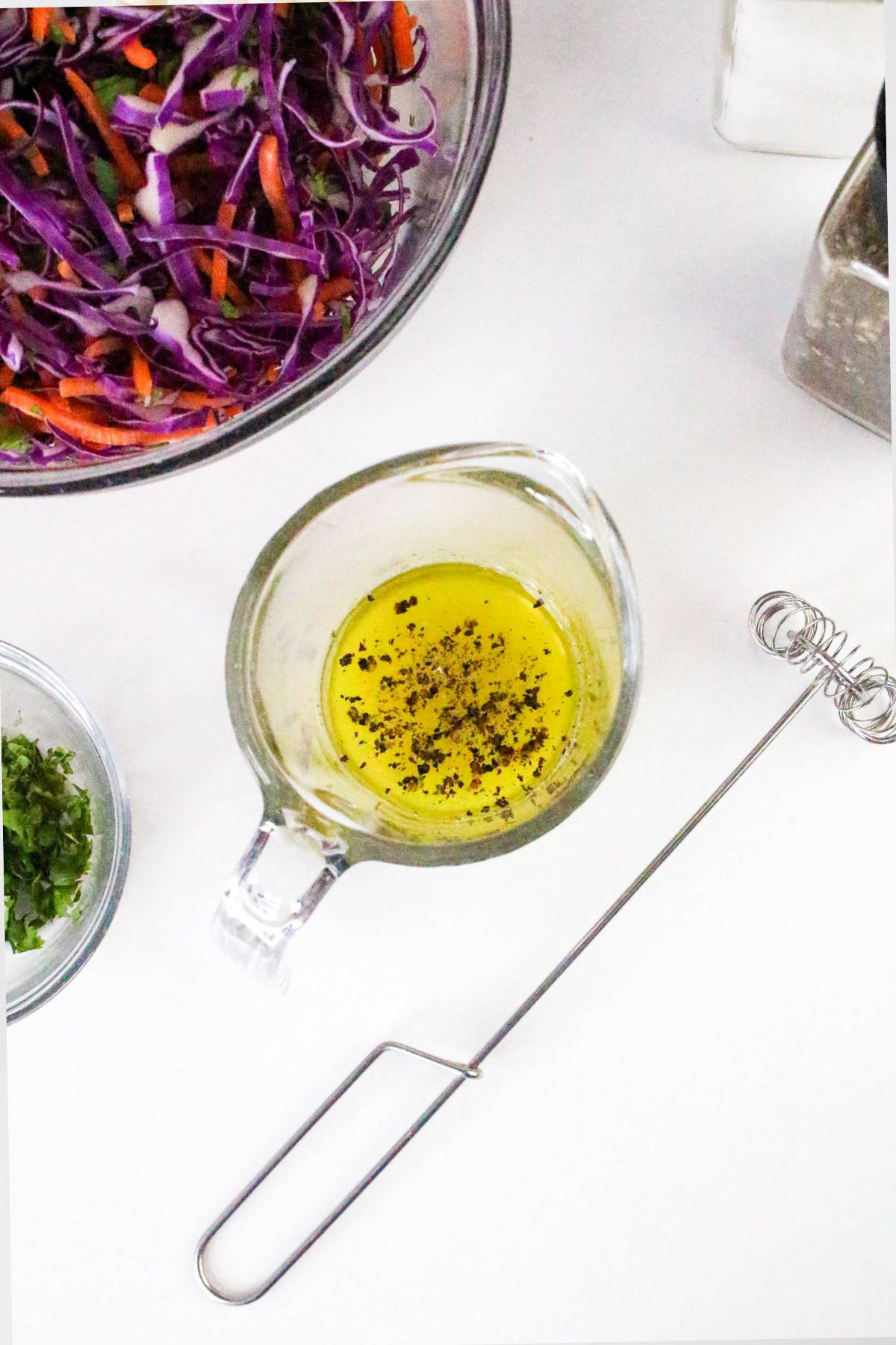 A glass measuring cup with oil and pepper, a handheld whisk, a bowl of chopped herbs, and a bowl of shredded purple cabbage and carrots on a white surface.