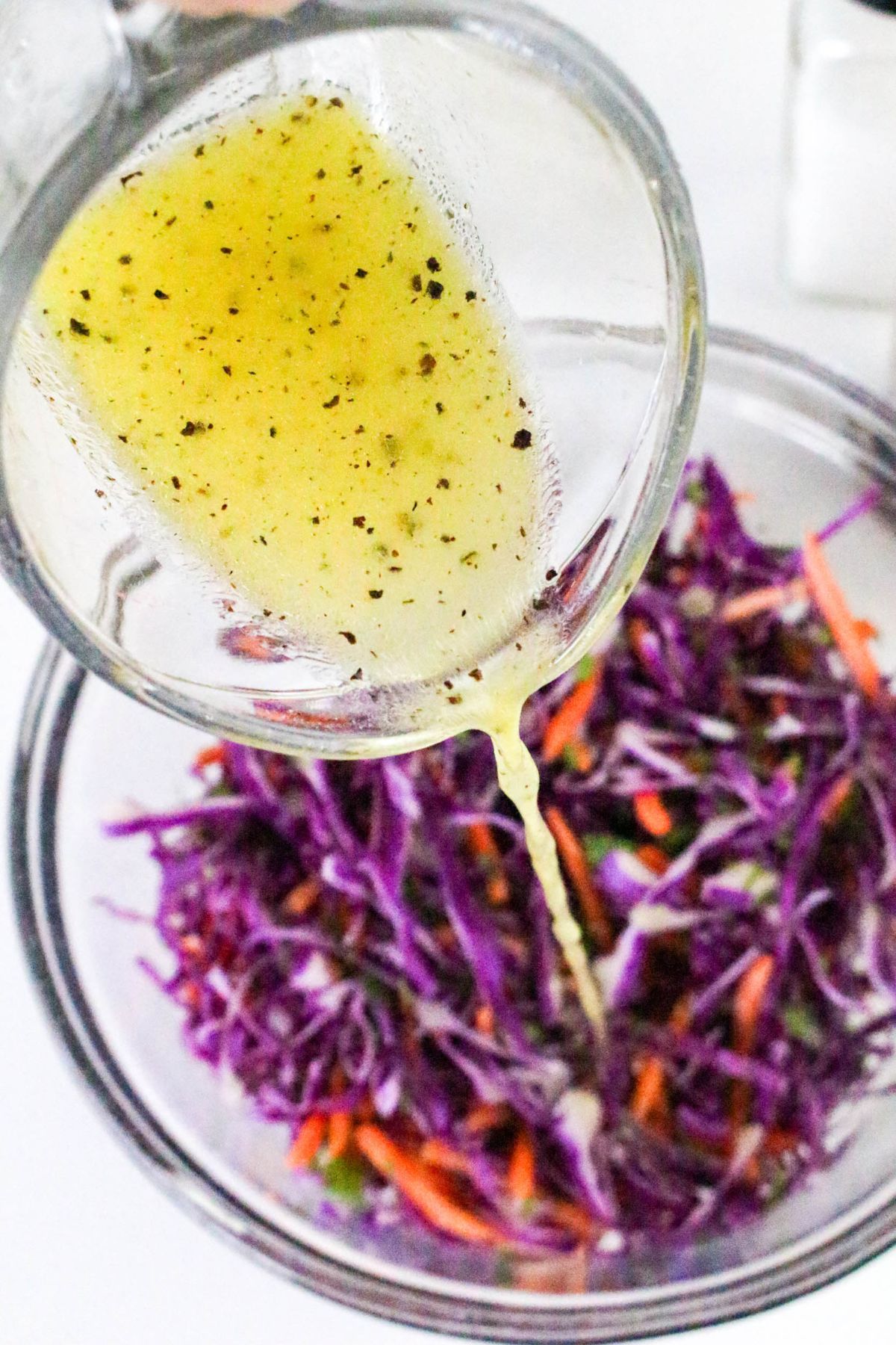 A close-up of mustard-colored vinaigrette being poured from a glass container onto a bowl of shredded purple cabbage and carrots.
