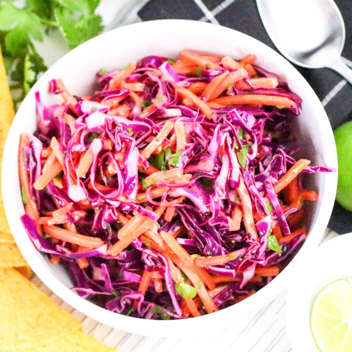 A white bowl filled with coleslaw made of shredded red cabbage, carrots, and green herbs sits on a striped surface. A spoon, lime, tortilla chips, and fresh cilantro are nearby.
