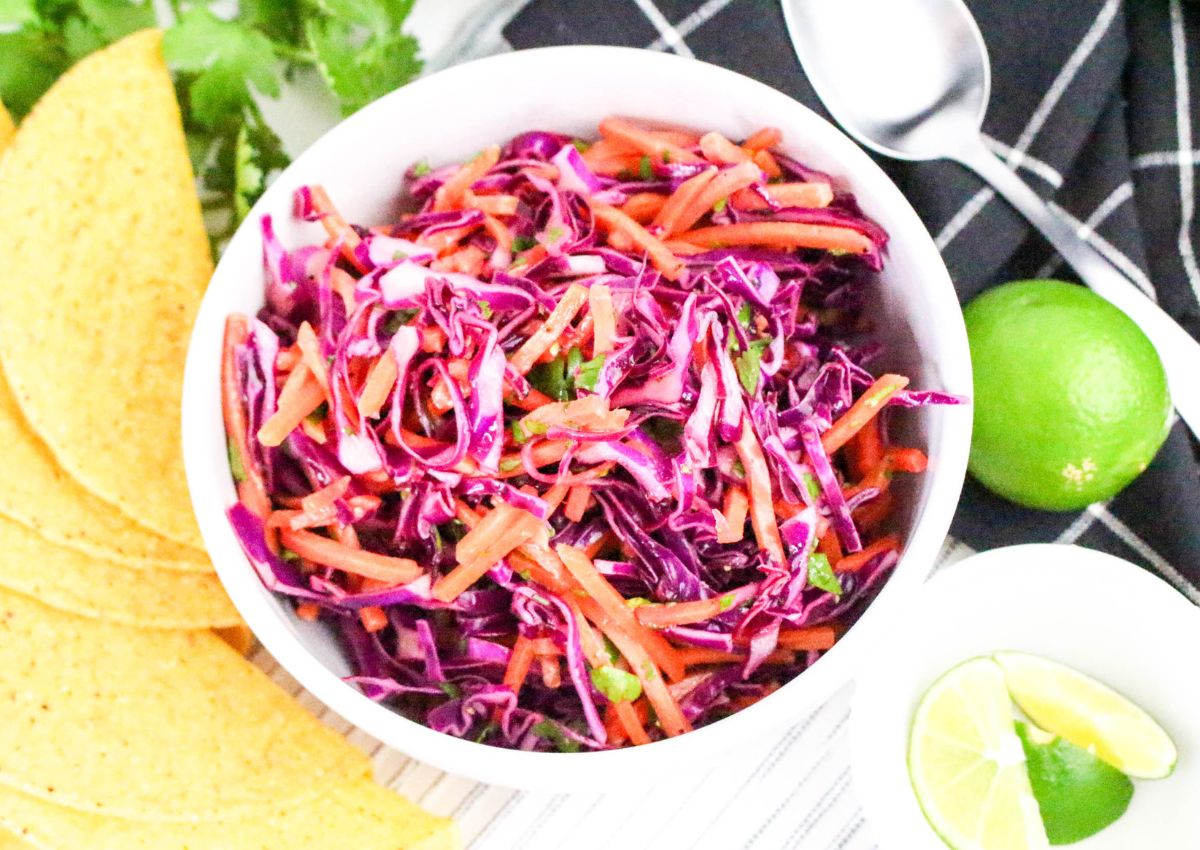 A white bowl filled with a colorful slaw made of shredded red cabbage, carrots, and cilantro sits next to taco shells, lime wedges, a whole lime, a spoon, and a black-and-white napkin.