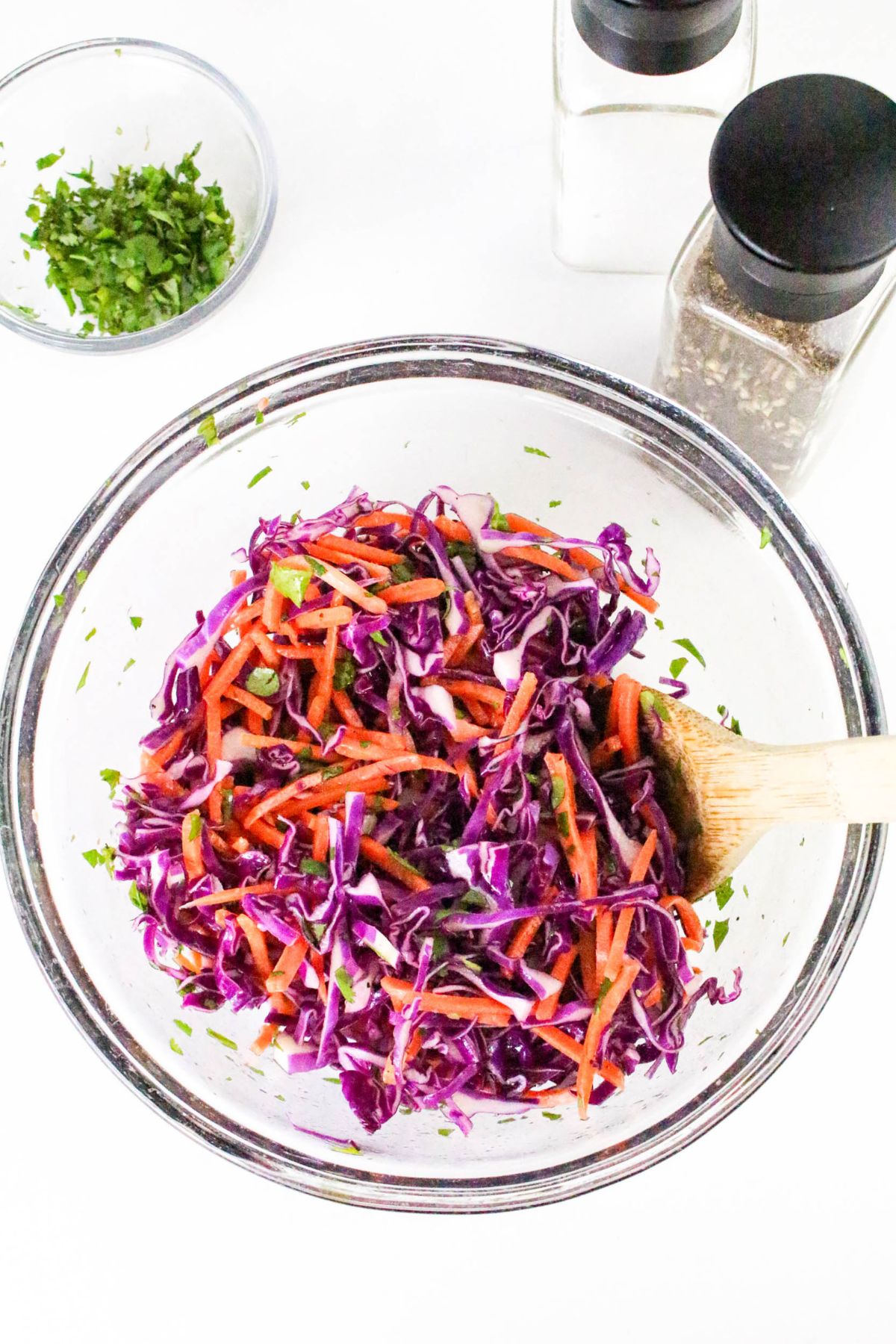 A glass bowl filled with shredded red cabbage, carrots, and green herbs sits on a white surface. A wooden spoon is in the bowl. Nearby are a small bowl of chopped herbs and two shakers for salt and pepper.