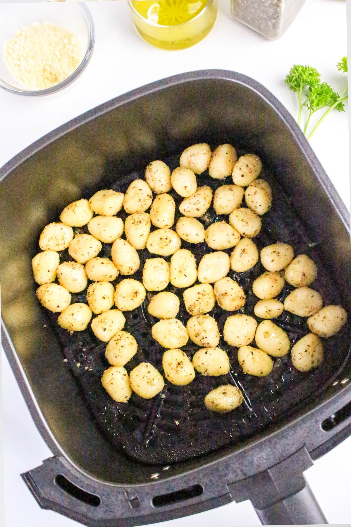 An air fryer basket filled with seasoned gnocchi, surrounded by small bowls of ingredients, a glass bottle of oil, a pepper grinder, and fresh parsley on a white surface.