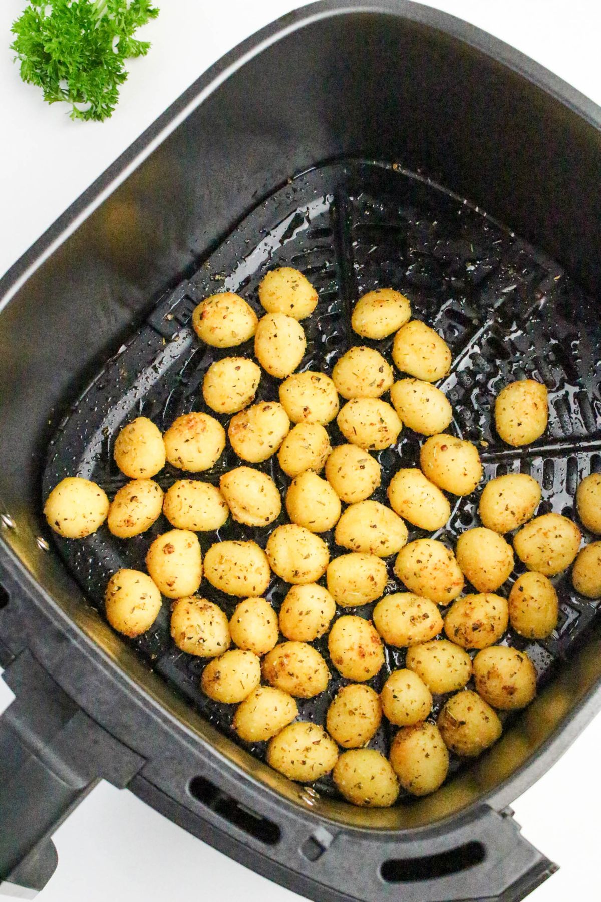 A black air fryer basket containing seasoned, cooked gnocchi pieces. A small sprig of parsley is visible in the top left corner on a white surface.