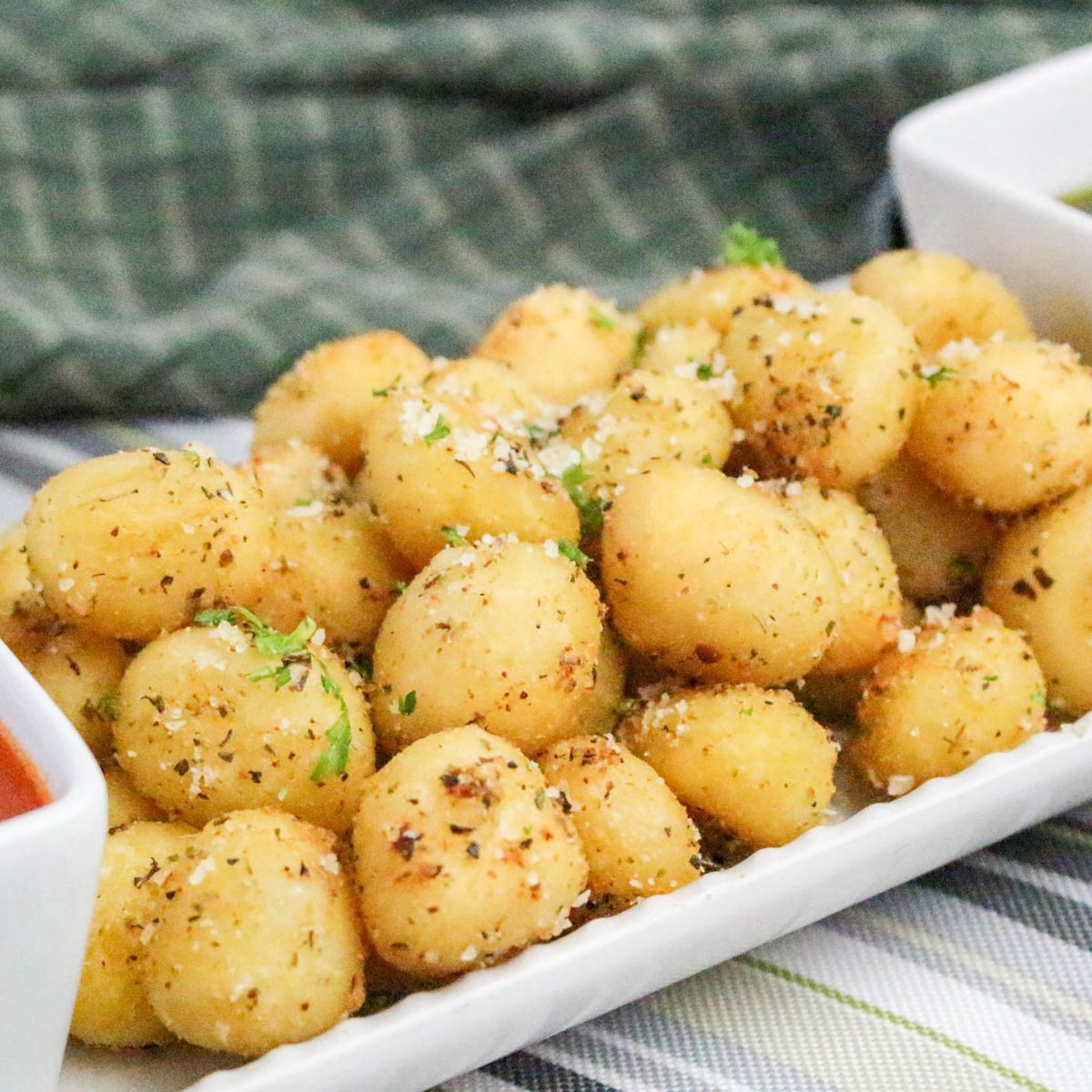 A rectangular white plate filled with seasoned, golden-brown gnocchi bites, garnished with herbs and grated cheese, with two dipping sauces in white bowls on either side. A green plaid cloth is in the background.
