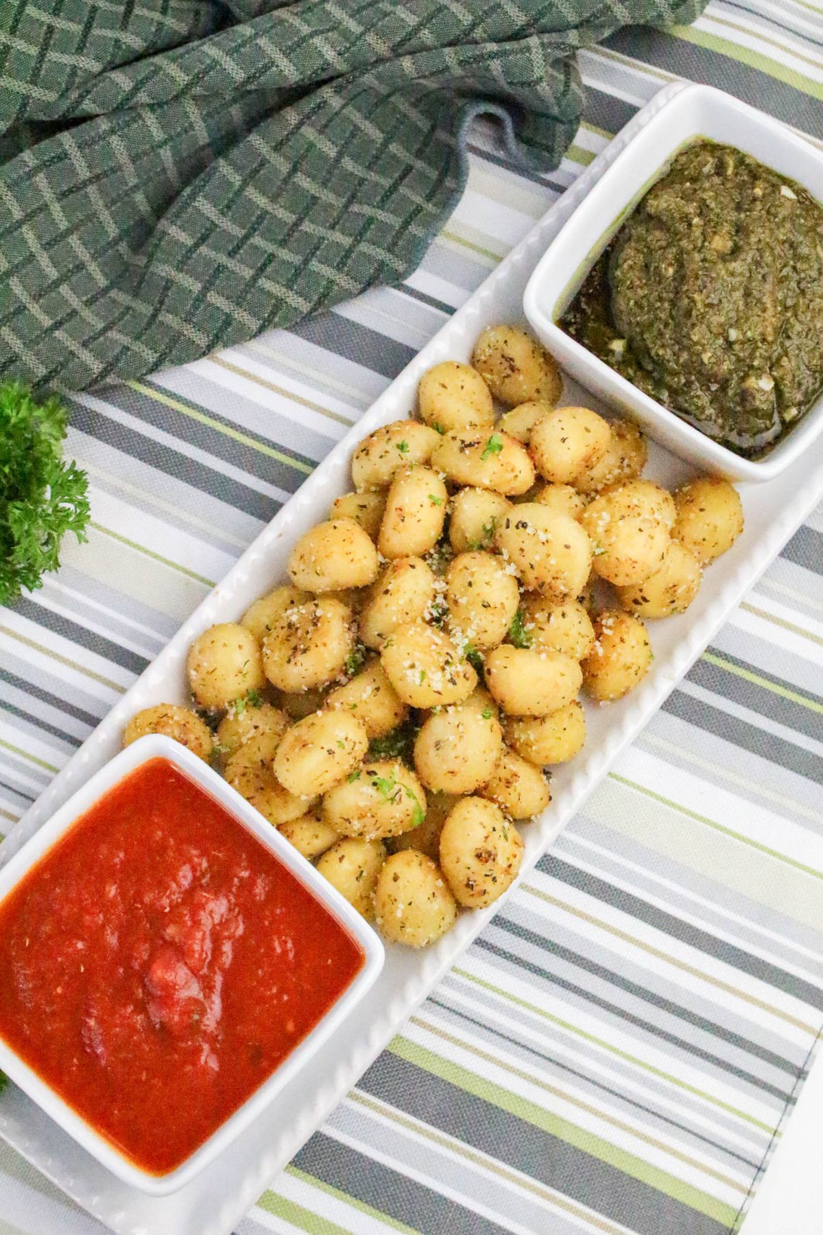 A rectangular white plate holds seasoned gnocchi in the center, flanked by two square dishes containing red marinara sauce on the left and green pesto sauce on the right. A green checked cloth is in the background.